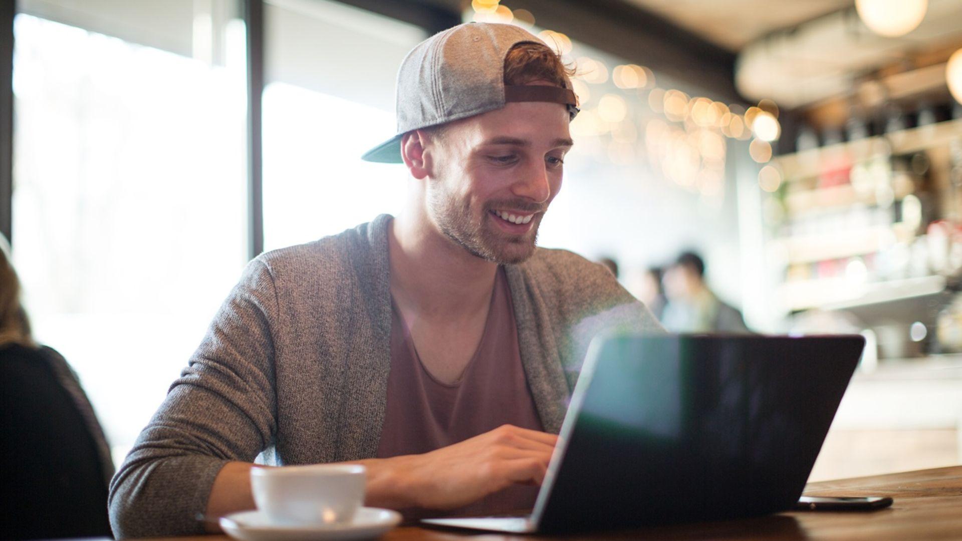 Junger Mann sitzt lächelnd vor dem Laptop und tippt etwas.