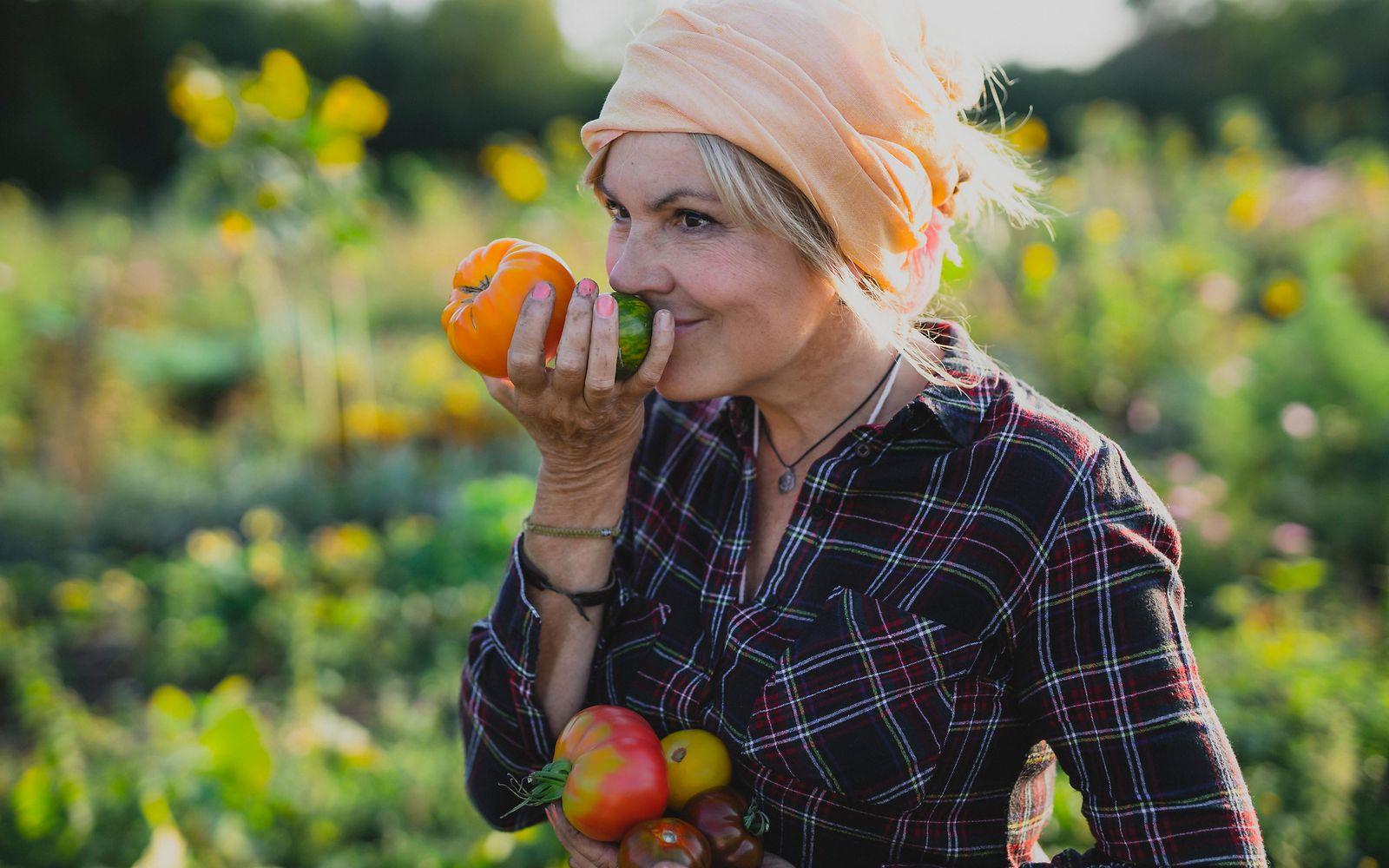 Wechseljahre und Bewerden: Eine Frau im mittleren Alter steht in einem Gemüsefeld und riecht an Tomaten