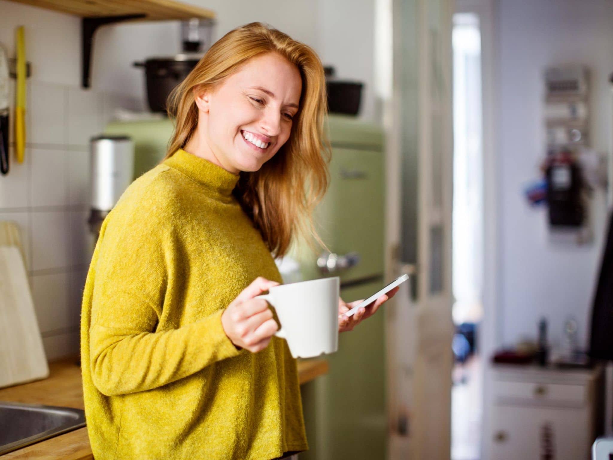 Kundenberatung: Junge Frau steht in der Küche und schaut fröhlich auf ihr Smartphone.