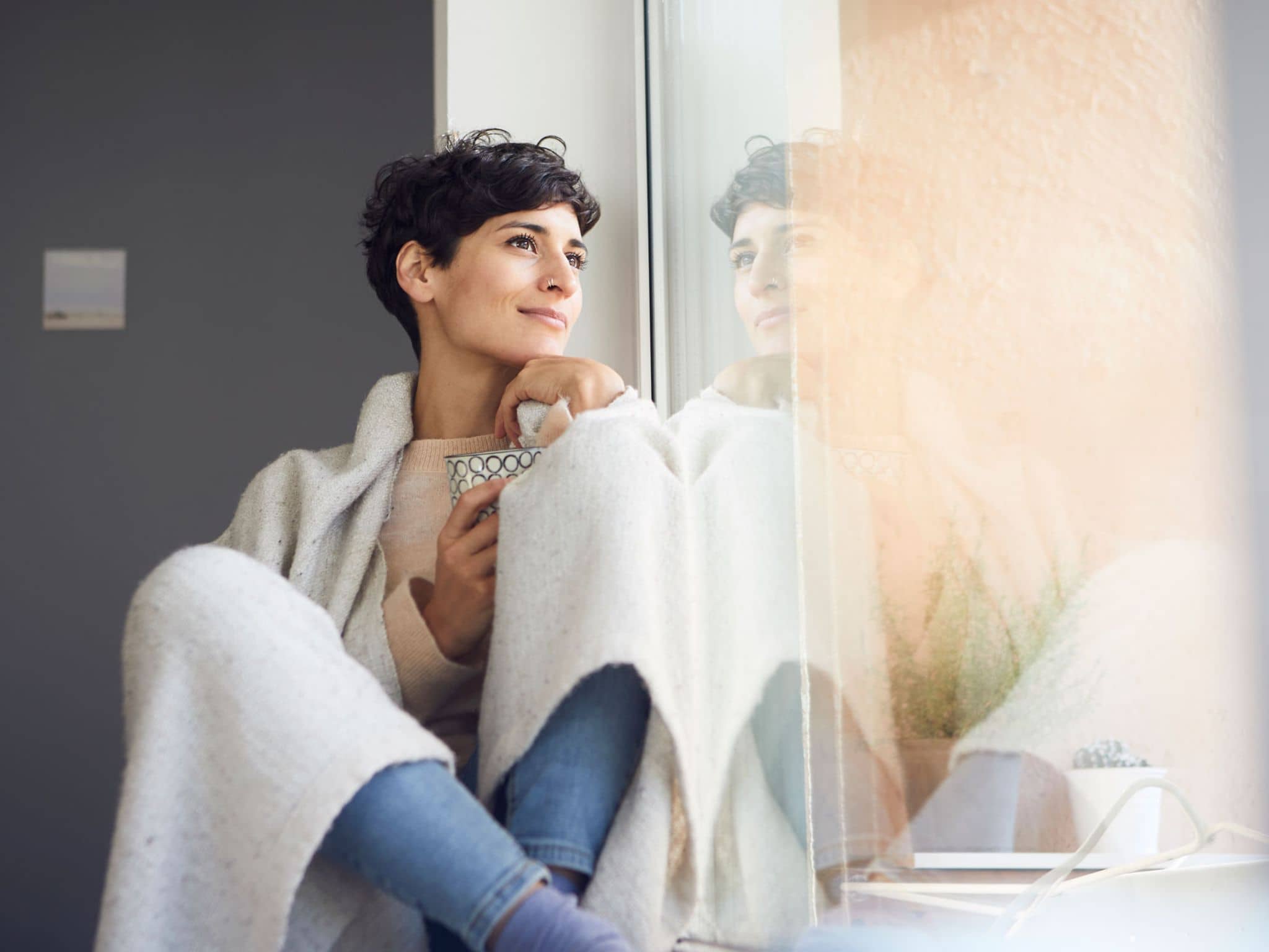 Krankengeld: Junge Frau sitzt in eine Wolldecke gehüllt am Fenster und schaut hinaus.