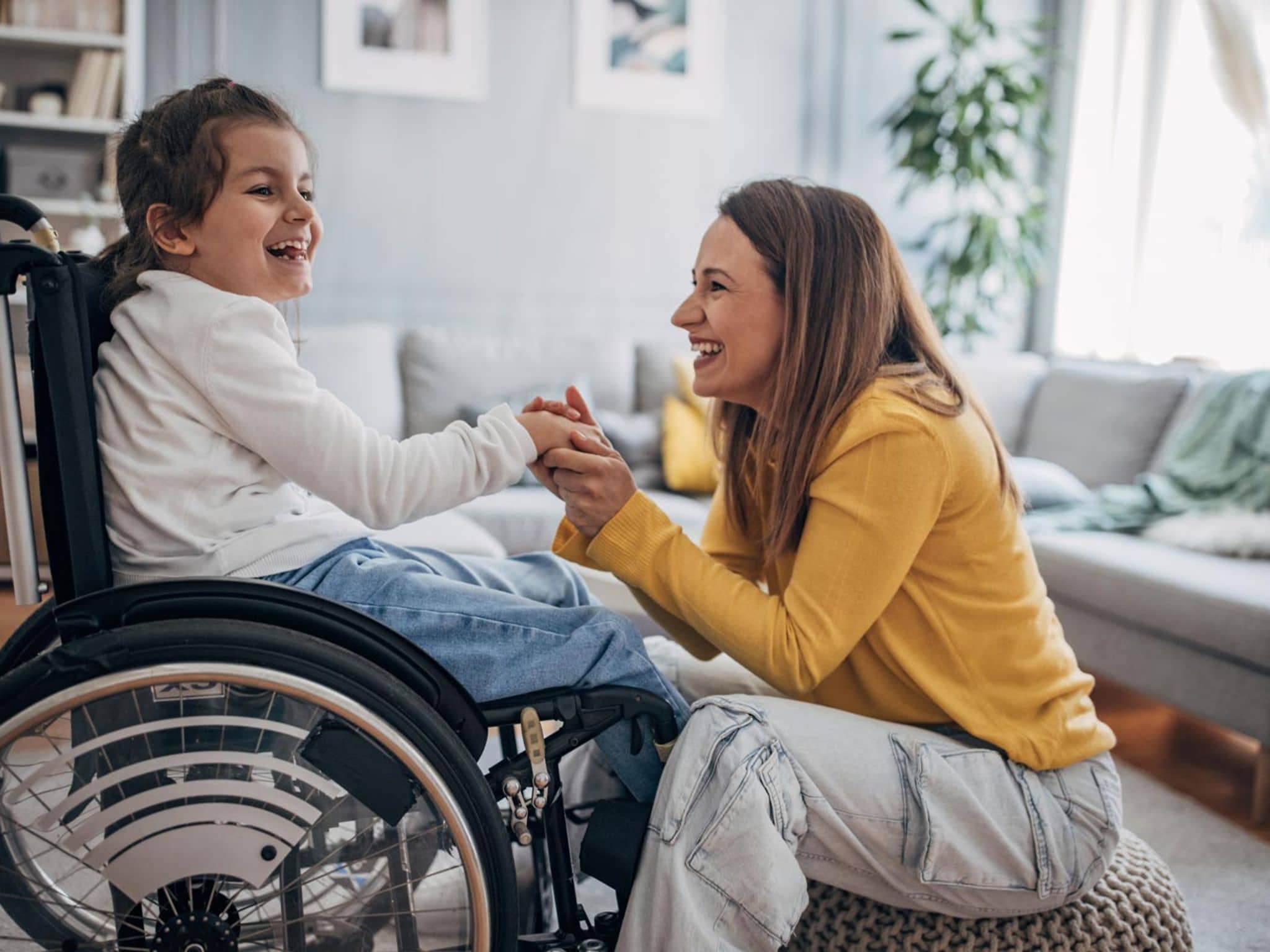 Hilfsmittel: Fröhliches Kind im Rollstuhl hält die Hände seiner Mutter, die im Wohnzimmer vor ihr auf einem Hocker sitzt.