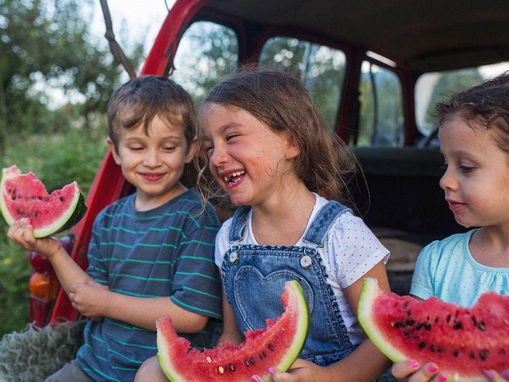 Safari-Kids: Drei Kinder sitzen lachend im offenen Kofferraum eines Autos und essen Melone.