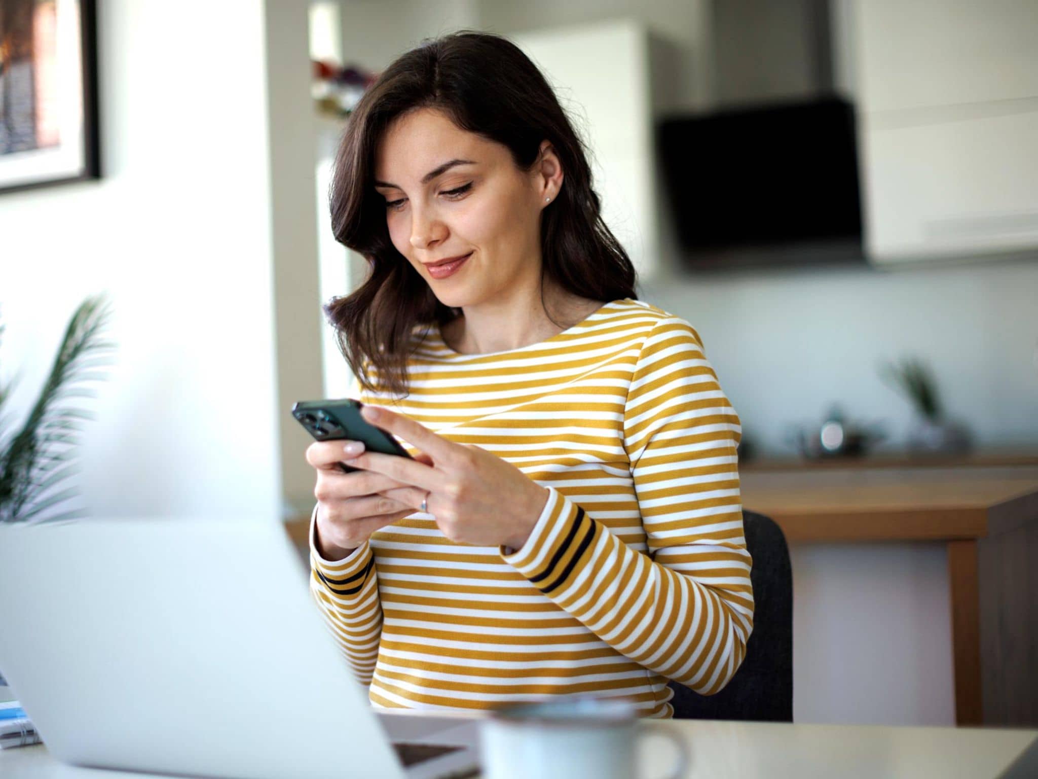 Lob & Beschwerde: Junge Frau sitzt zuhause am Tisch und tippt auf ihrem Smartphone.