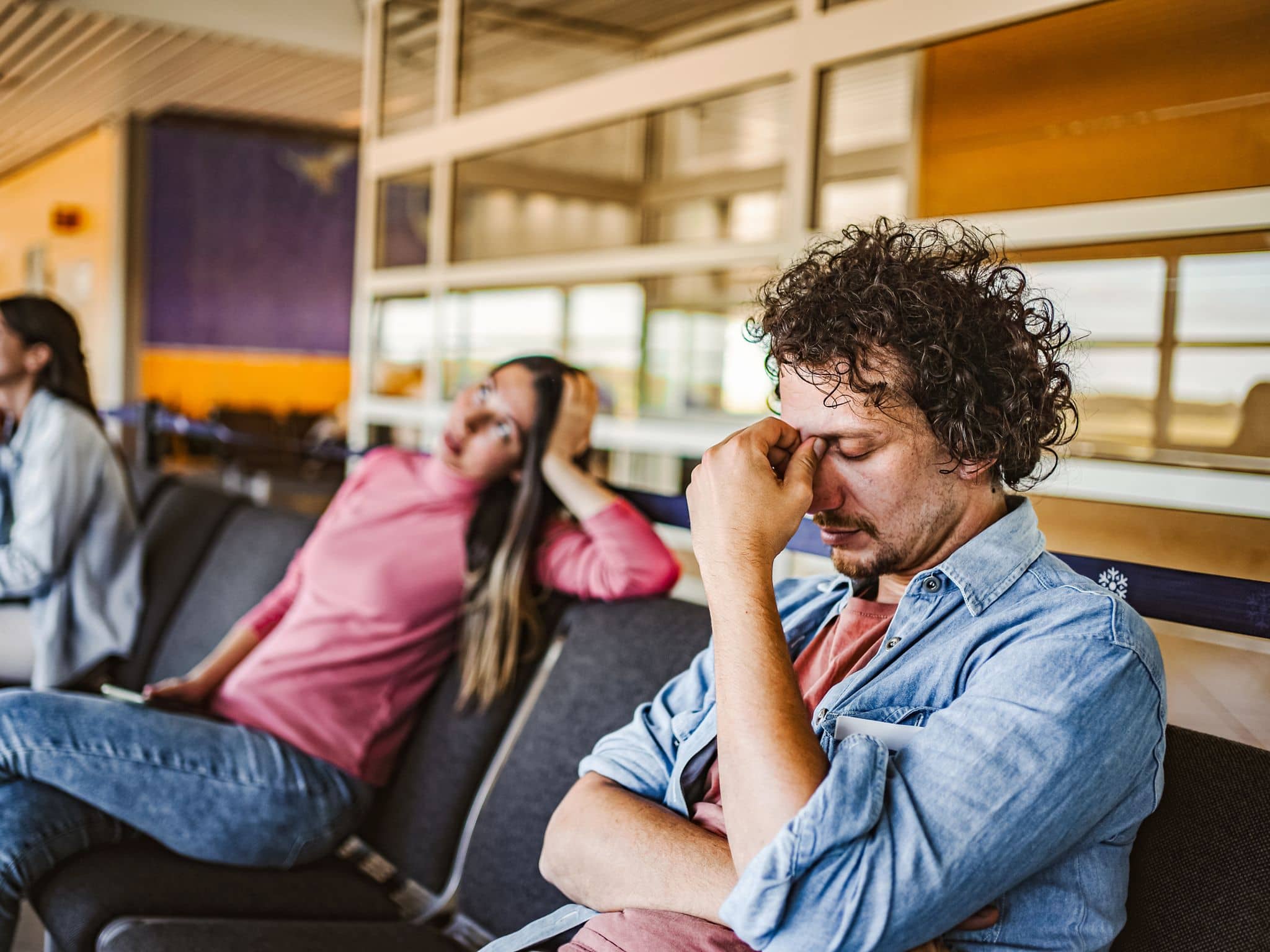 Mann am Flughafen sitzt mit geschlossenen Augen. Im Hintergrund zwei sitzende Frauen.