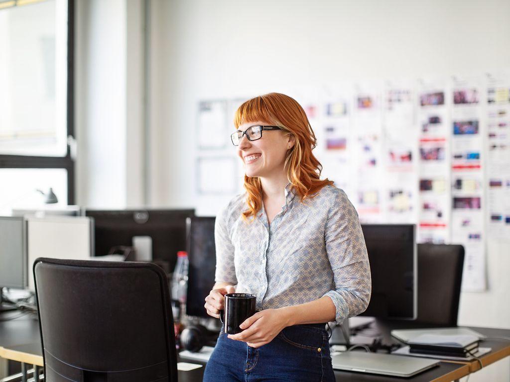 Bild: Frau mit roten Haaren steht im Büro mit einer Kaffeetasse