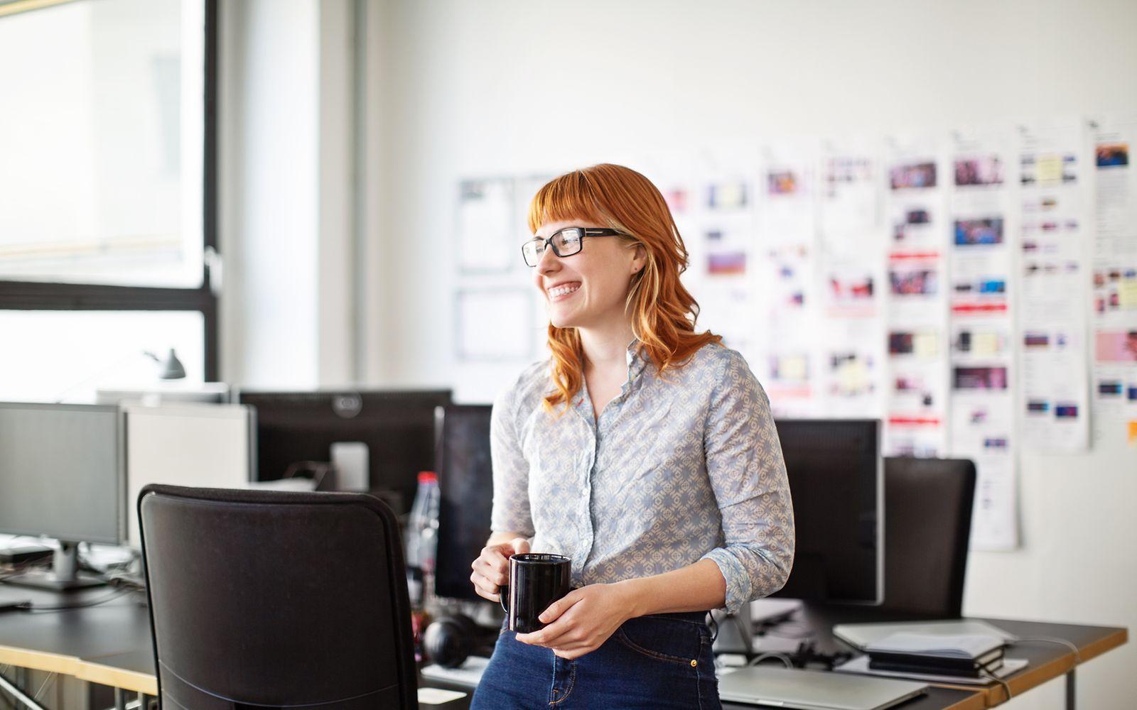 Bild: Frau mit roten Haaren steht im Büro mit einer Kaffeetasse