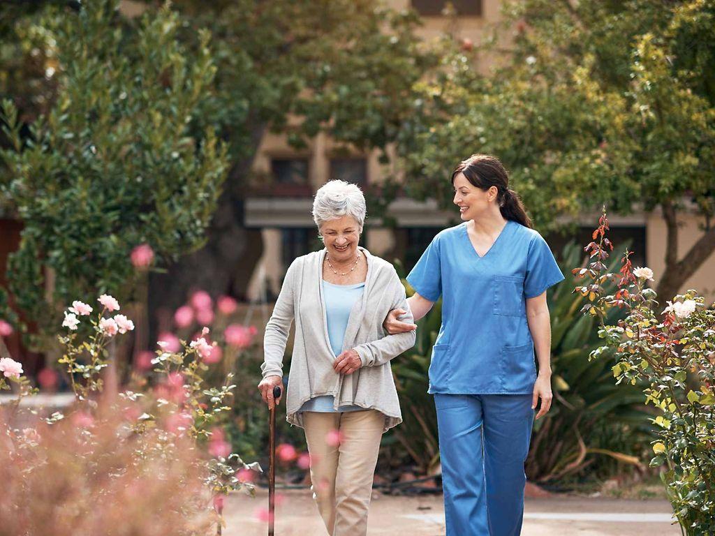 Bild: Pflegerin und Patientin gehen im Rosengarten spazieren.