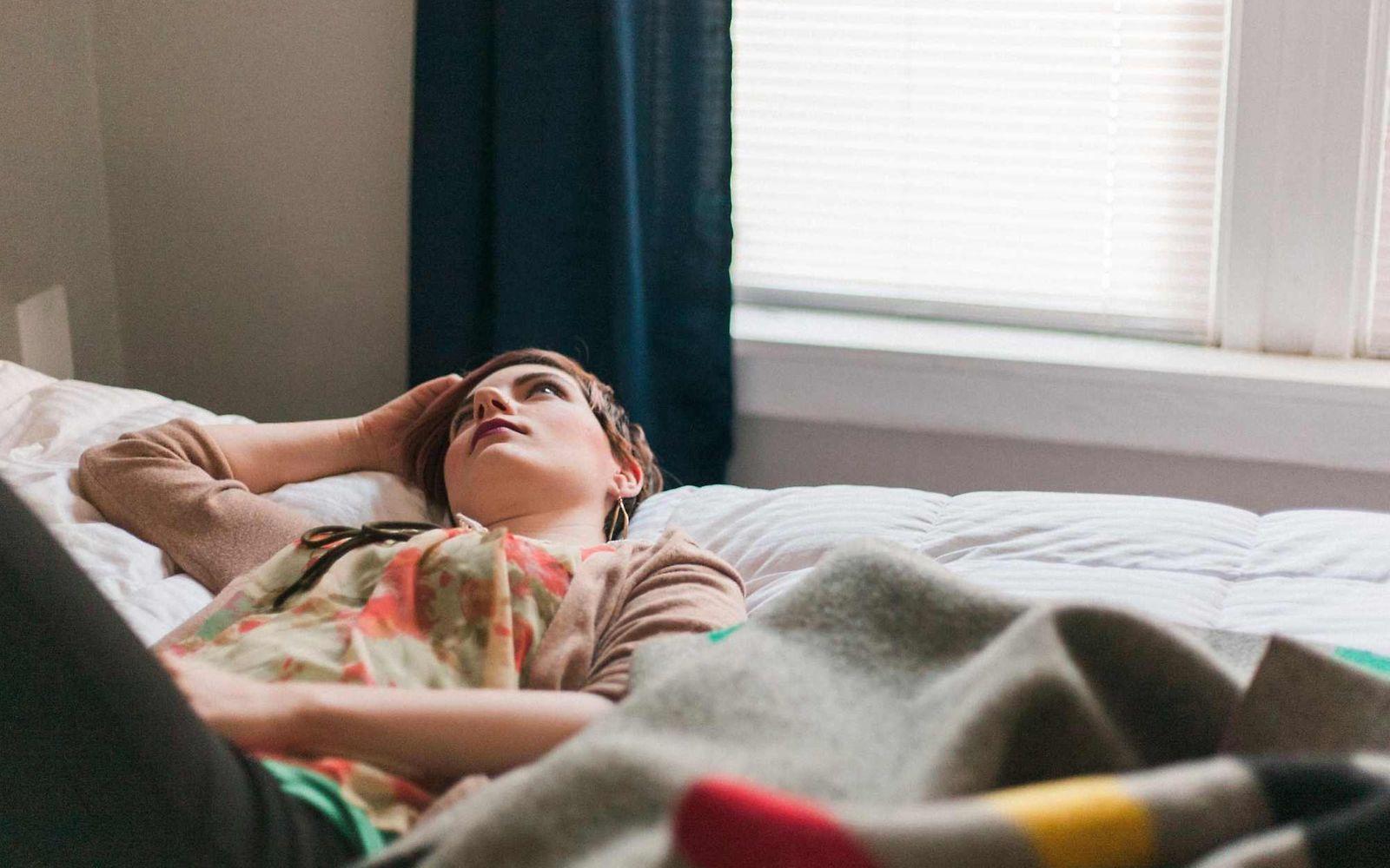 Migräne: Junge Frau liegt auf dem Bett, hält die Hand an den Kopf und guckt zur Zimmerdecke.