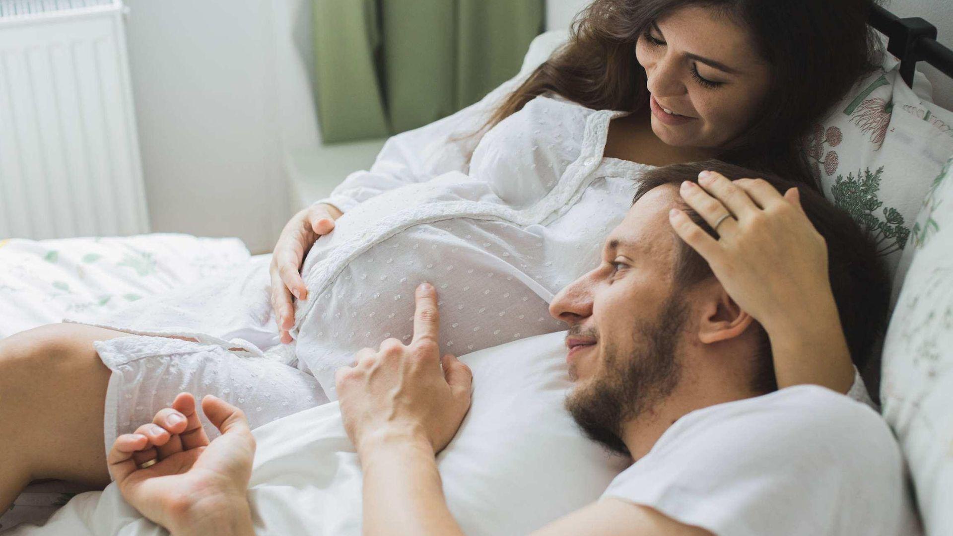 Schwangerschaft: Schwangere Frau liegt mit Ihrem Partner im Bett und streicht sich über den Bauch.