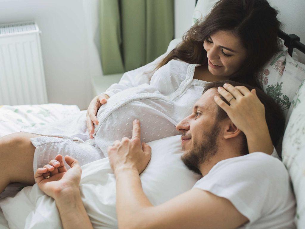 Schwangerschaft: Schwangere Frau liegt mit Ihrem Partner im Bett und streicht sich über den Bauch.