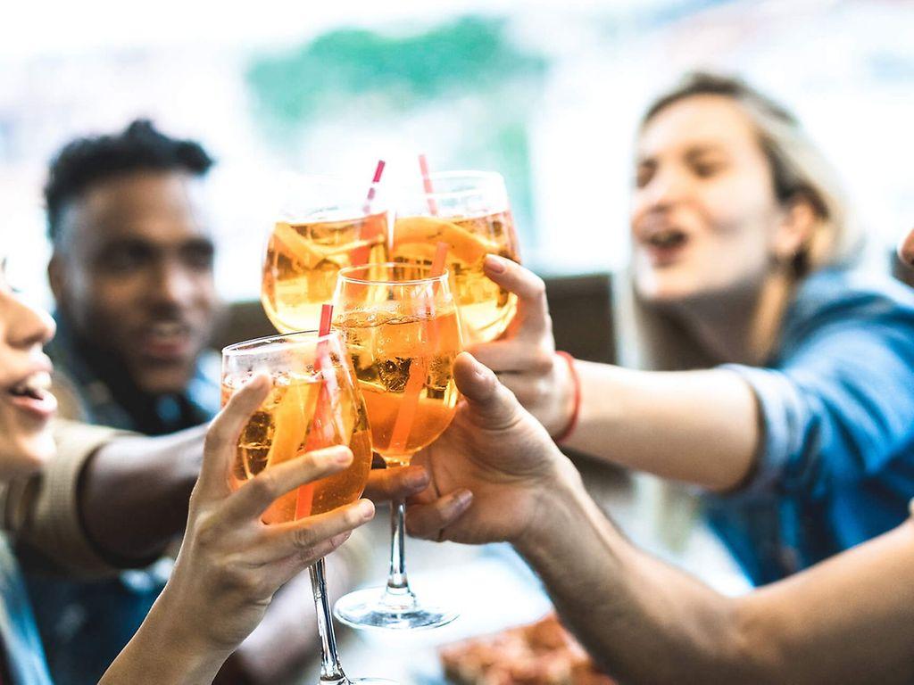 Alkohol Droge Nr.1: Vier junge Leute sitzen zusammen und stoßen freudig mit einem Aperol-Cocktail an