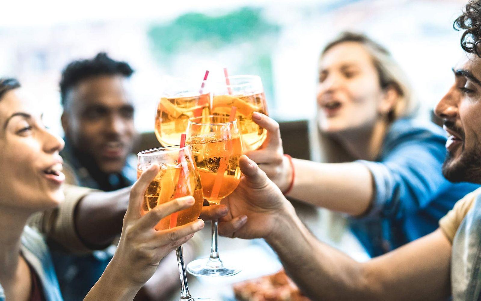 Alkohol Droge Nr.1: Vier junge Leute sitzen zusammen und stoßen freudig mit einem Aperol-Cocktail an