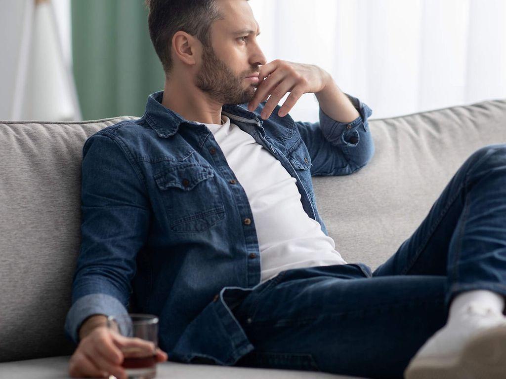 Alkoholsüchtig?: Mann sitzt nachdenklich auf dem Sofa mit einem alkoholischen Drink in der Hand