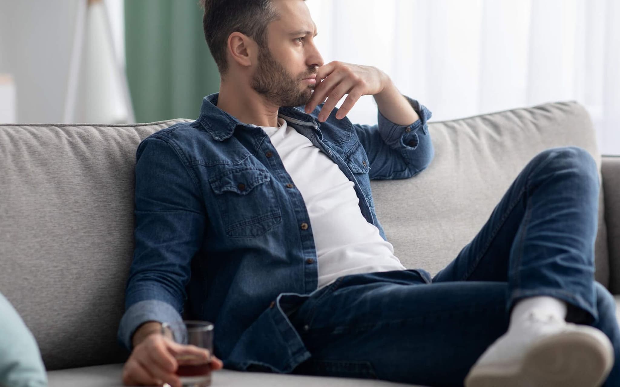 Alkoholsüchtig?: Mann sitzt nachdenklich auf dem Sofa mit einem alkoholischen Drink in der Hand