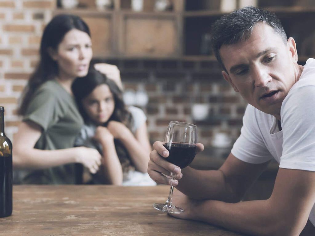 Alkoholismus für Angehörige: Mann trinkt Alkohol und im Hintergrund sitzt seine vorwurfsvolle Frau und ein kleines Mädchen.