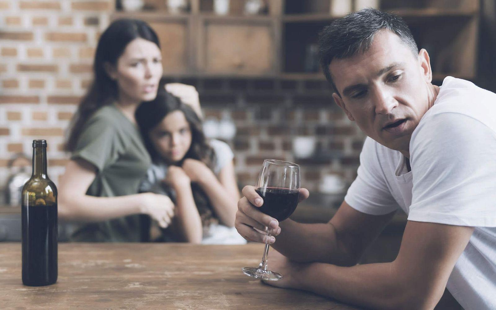Alkoholismus für Angehörige: Mann trinkt Alkohol und im Hintergrund sitzt seine vorwurfsvolle Frau und ein kleines Mädchen.