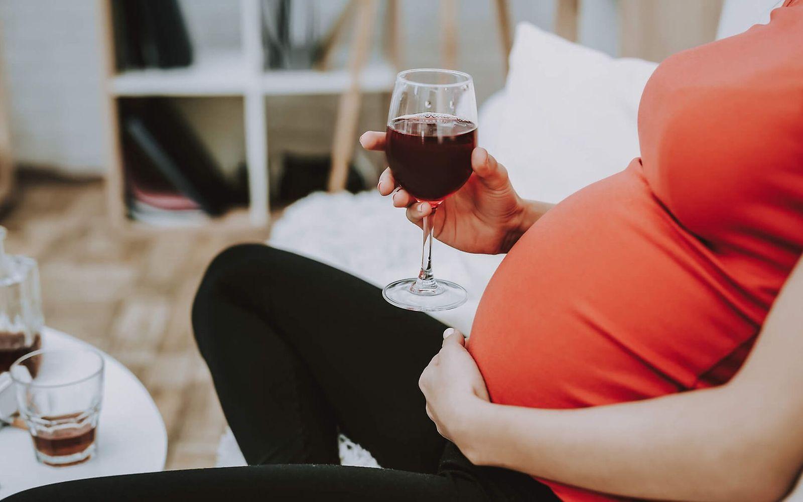 Alkohol in der Schwangerschaft: Schwangere sitzt vor einem Tisch mit Flaschen und Zigaretten und hält ein Glas Wein in der Hand 