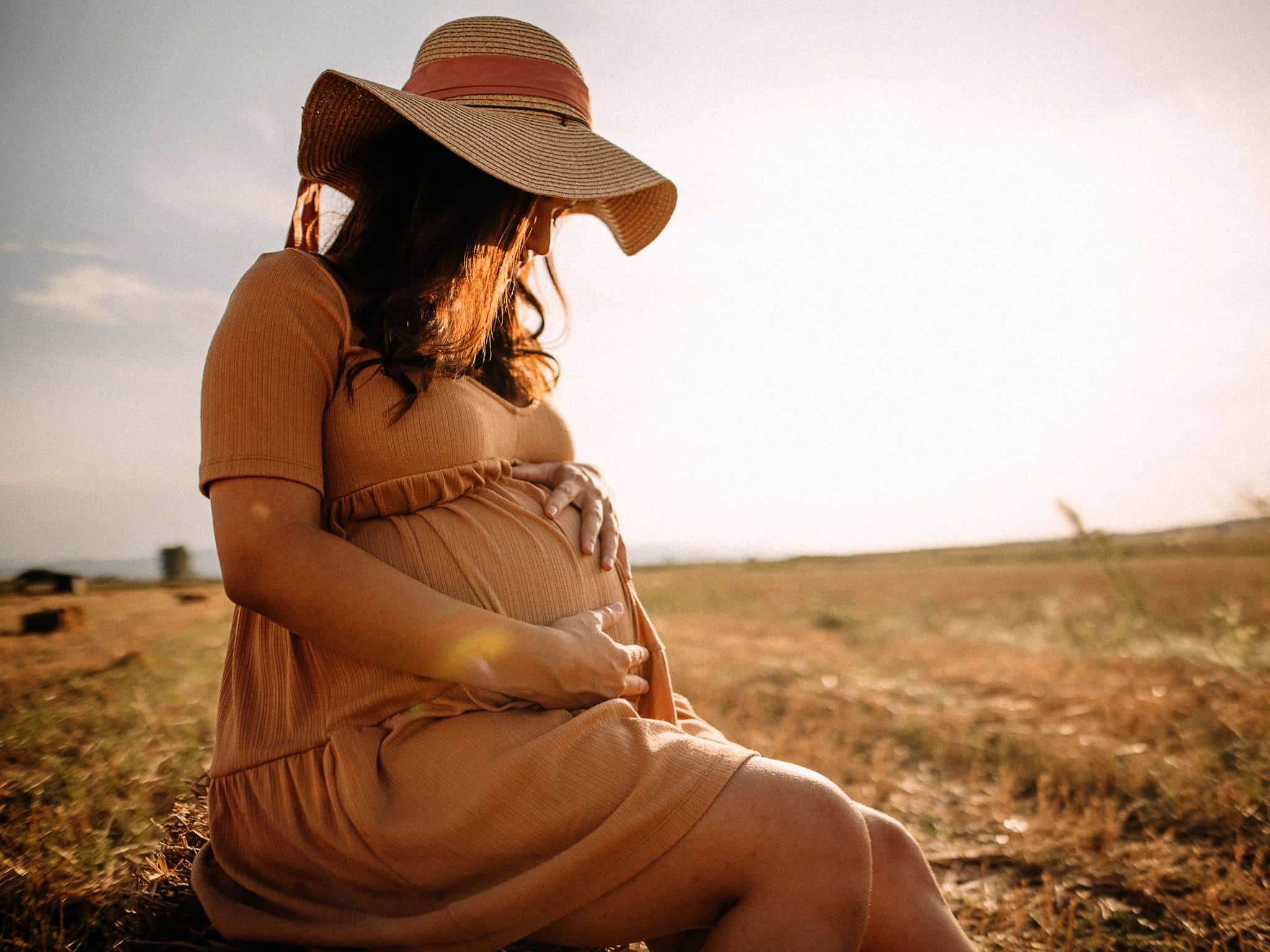 Frau mit luftigem Kleid und Sonnenhut sitzt auf einer Wiese und betrachtet ihren Babybauch.