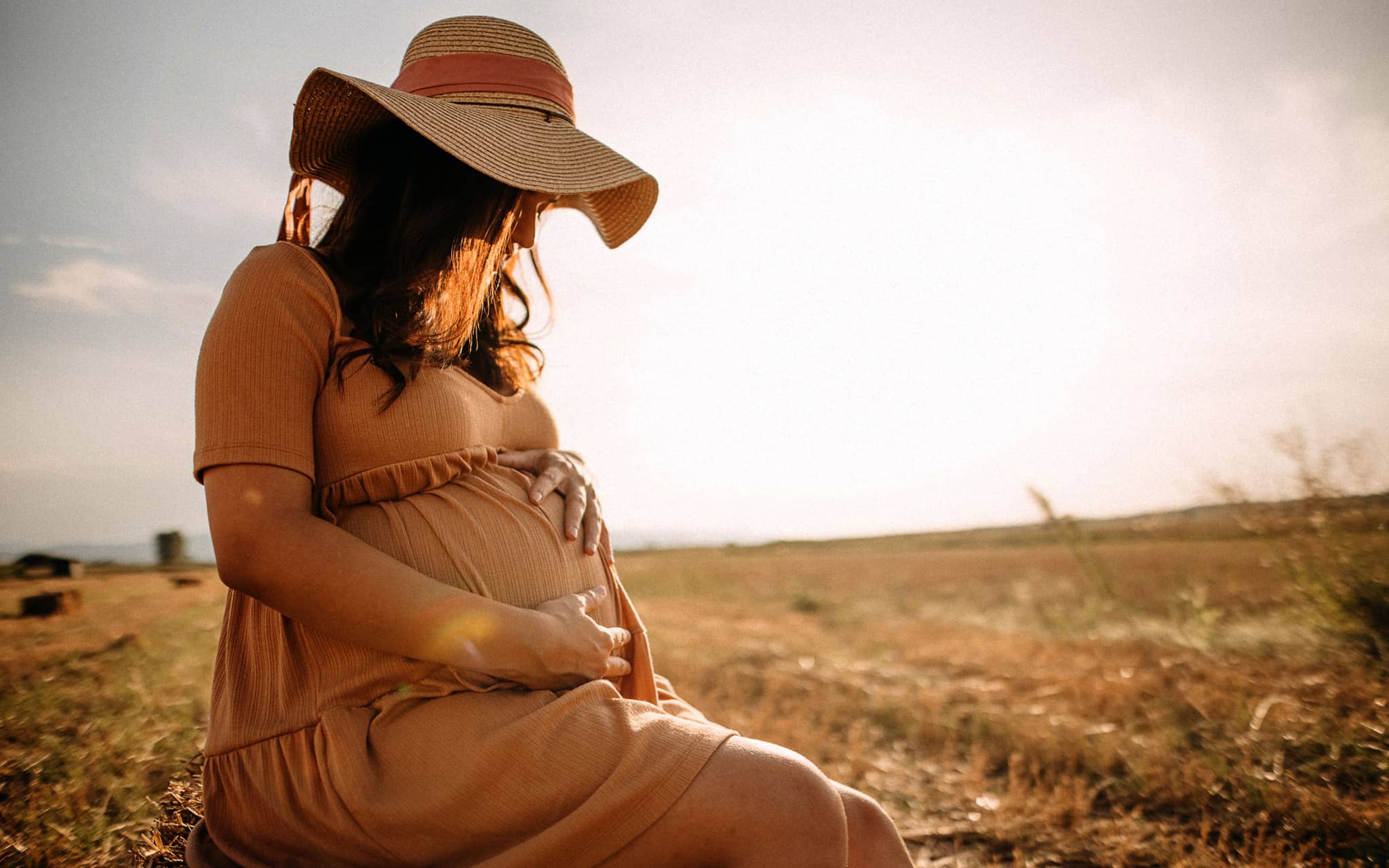 Frau mit luftigem Kleid und Sonnenhut sitzt auf einer Wiese und betrachtet ihren Babybauch.