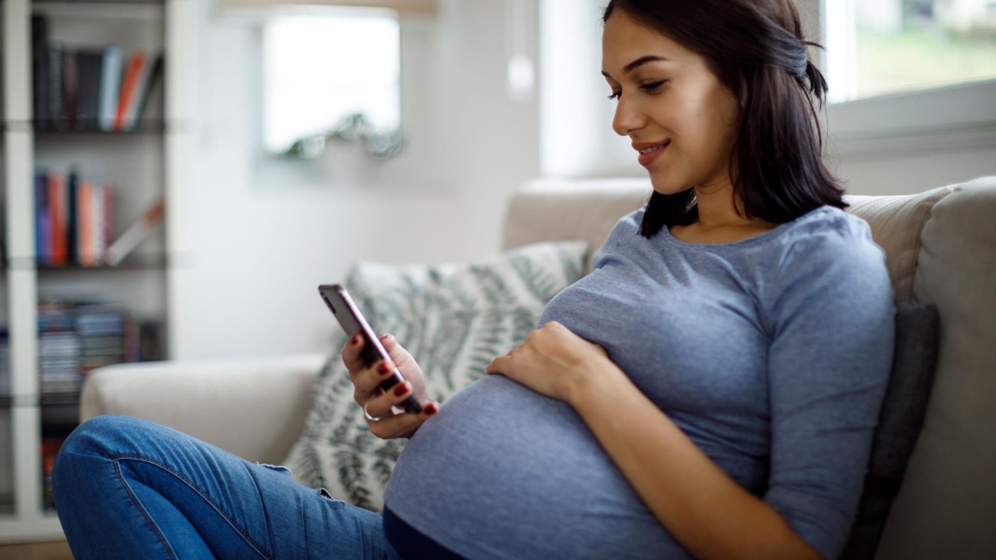 Mutterschaftsgeld: Lächelnde schwangere Frau sitzt zu Hause auf dem Sofa und schaut auf ihr Smartphone.