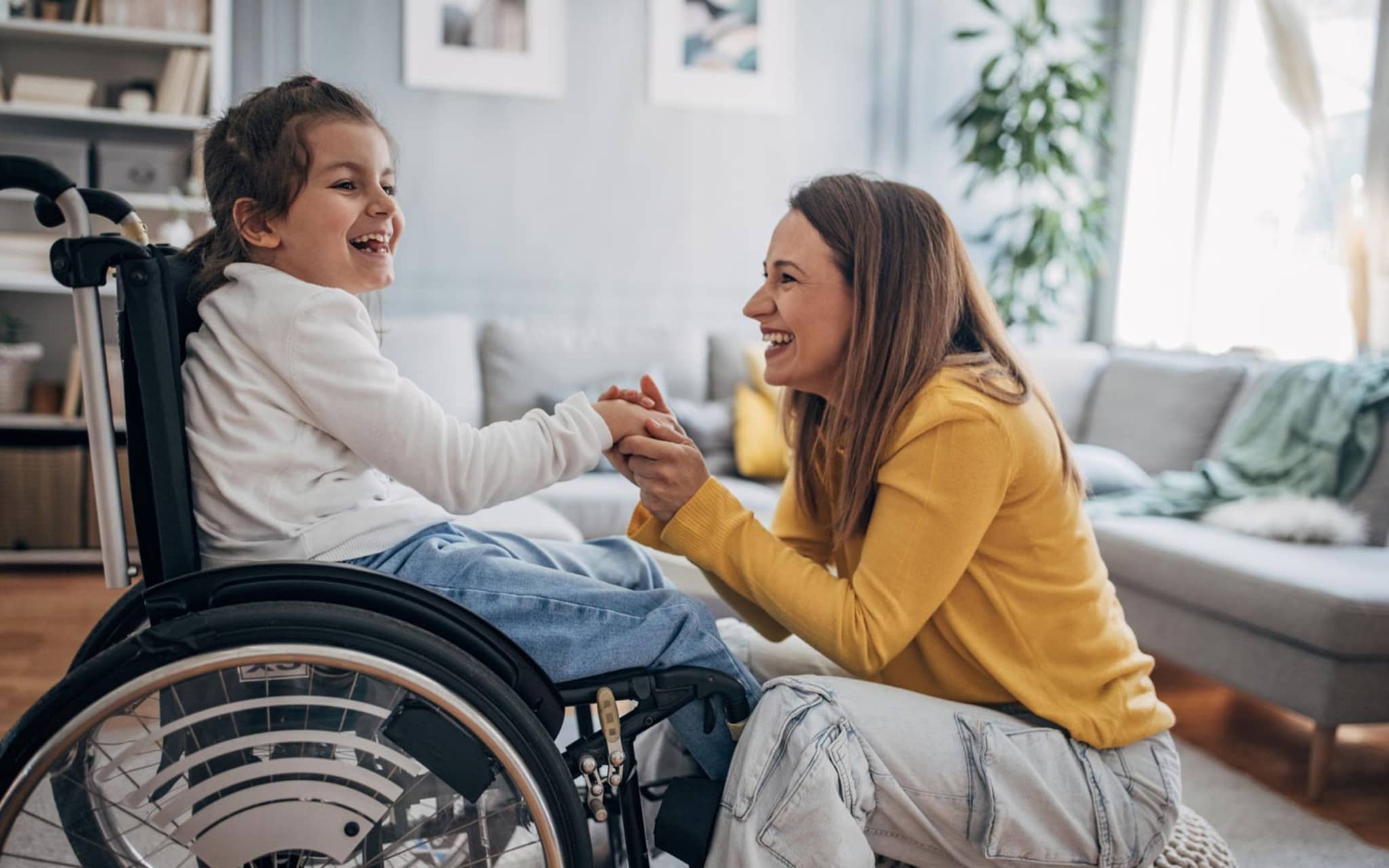 Hilfsmittel: Fröhliches Kind im Rollstuhl hält die Hände seiner Mutter, die im Wohnzimmer vor ihr auf einem Hocker sitzt.