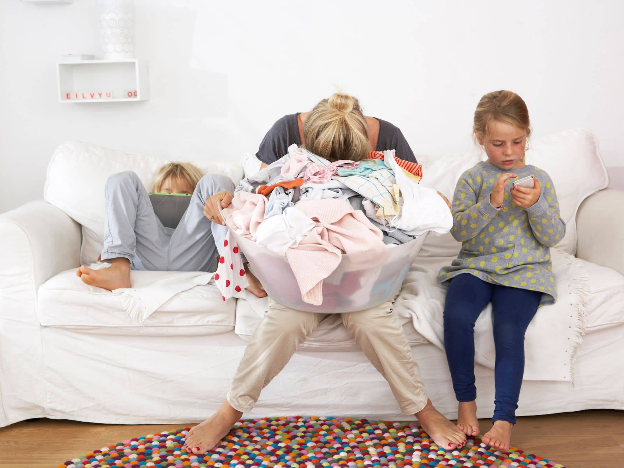 Mental Load: Eine Mutter sitzt auf einem Sofa und hält ihren Kopf in den Händen, zwei Kinder toben neben ihr