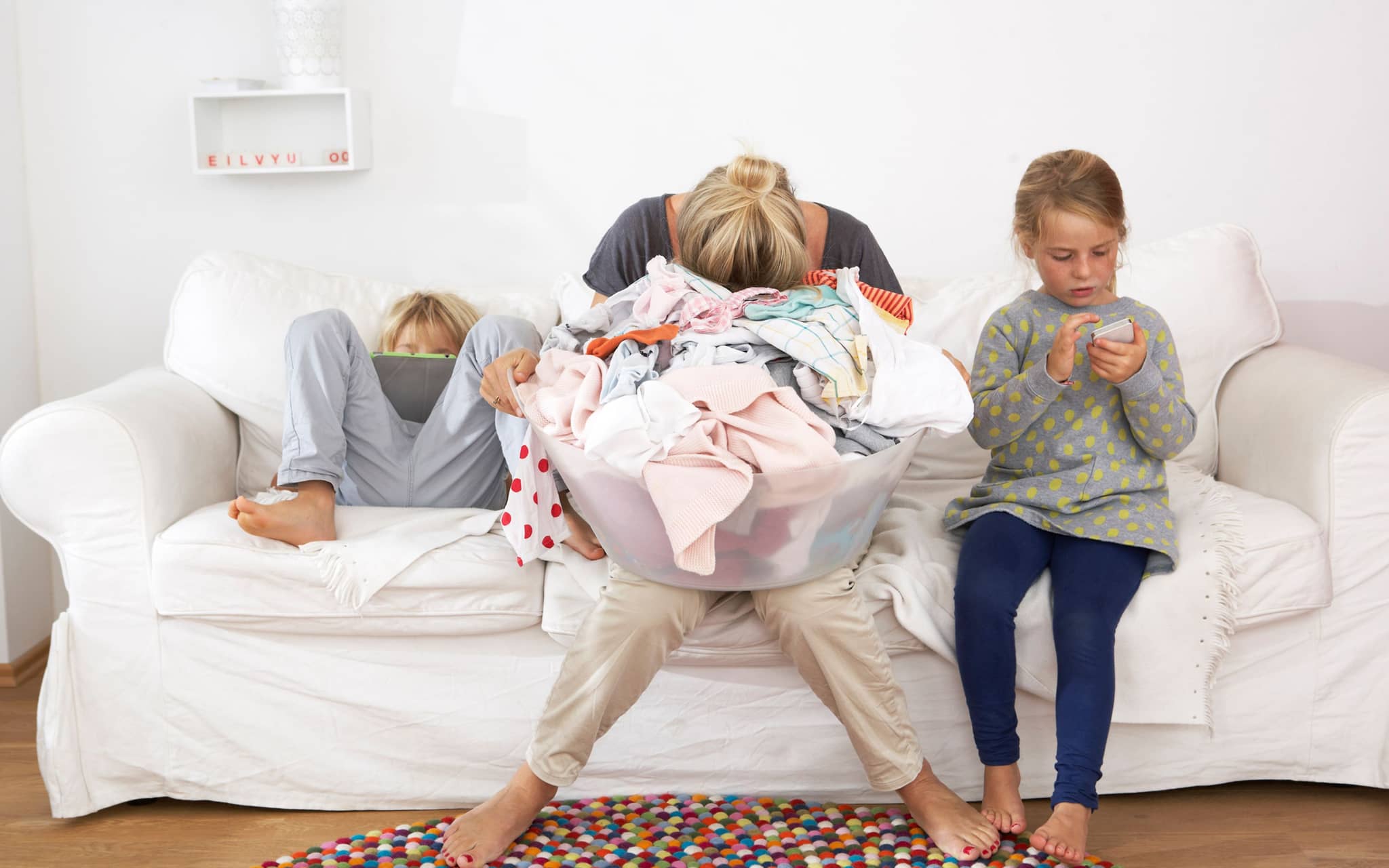 Mental Load: Eine Mutter sitzt auf einem Sofa und hält ihren Kopf in den Händen, zwei Kinder toben neben ihr