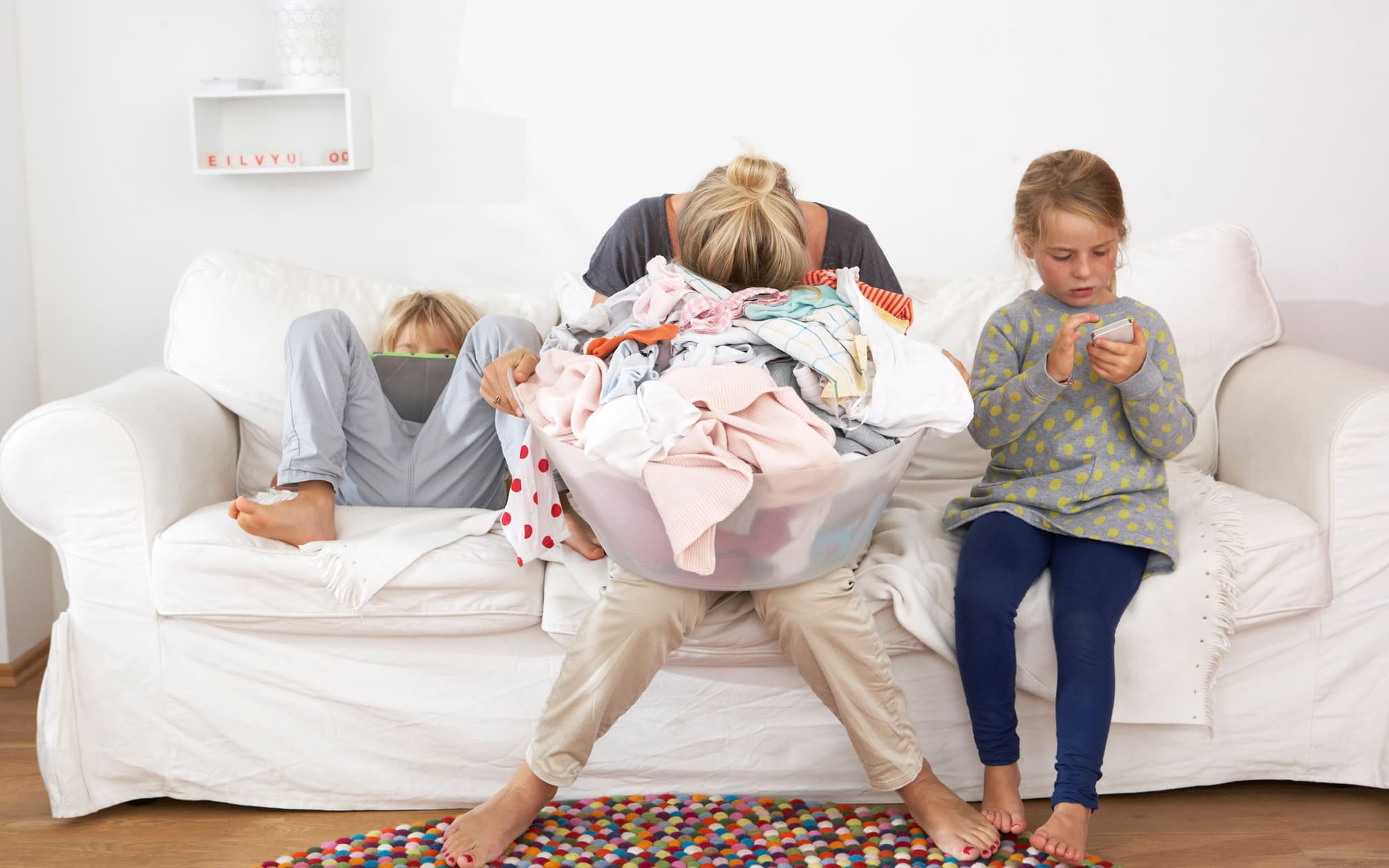 Mental Load: Eine Mutter sitzt auf einem Sofa und hält ihren Kopf in den Händen, zwei Kinder toben neben ihr