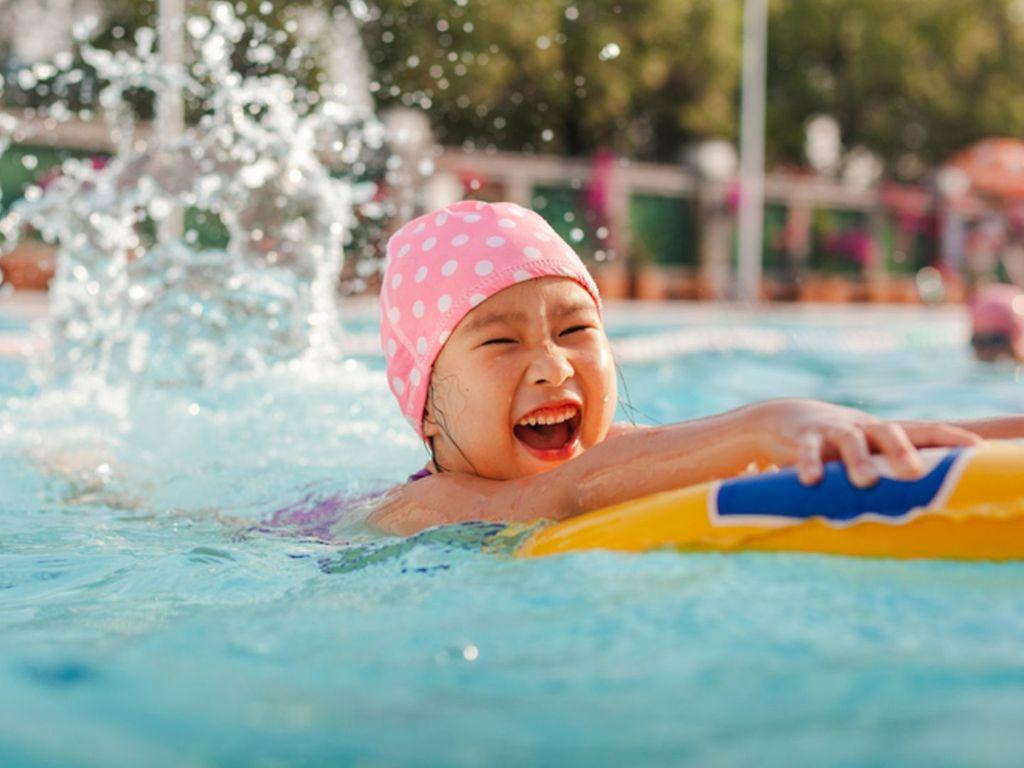 Mit Kindern schwimmen lernen: Kleines fröhliches Mädchen schwimmt mit einem Schwimmring und Badekappe im Pool.