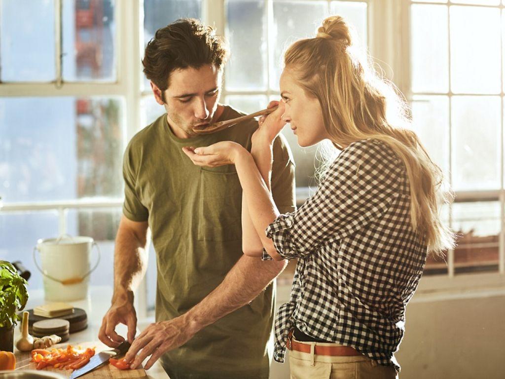 Ernährungskurse: Frau hält Mann Kochlöffel vor den Mund, damit er das Essen probiert.