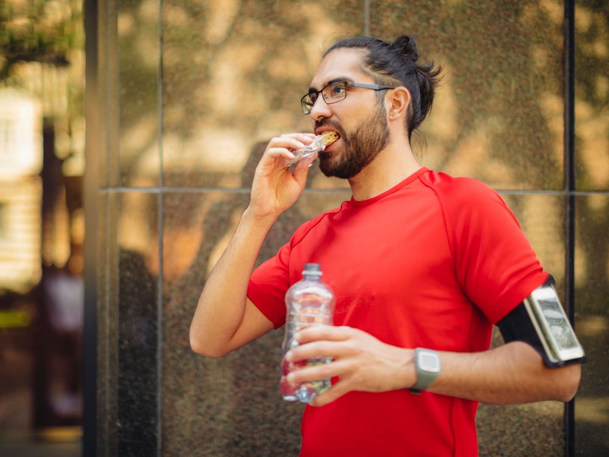Fruchtriegel nach dem Sport: Mann isst nach dem Joggen einen Snackriegel und hält eine Flasche Wasser in der Hand.