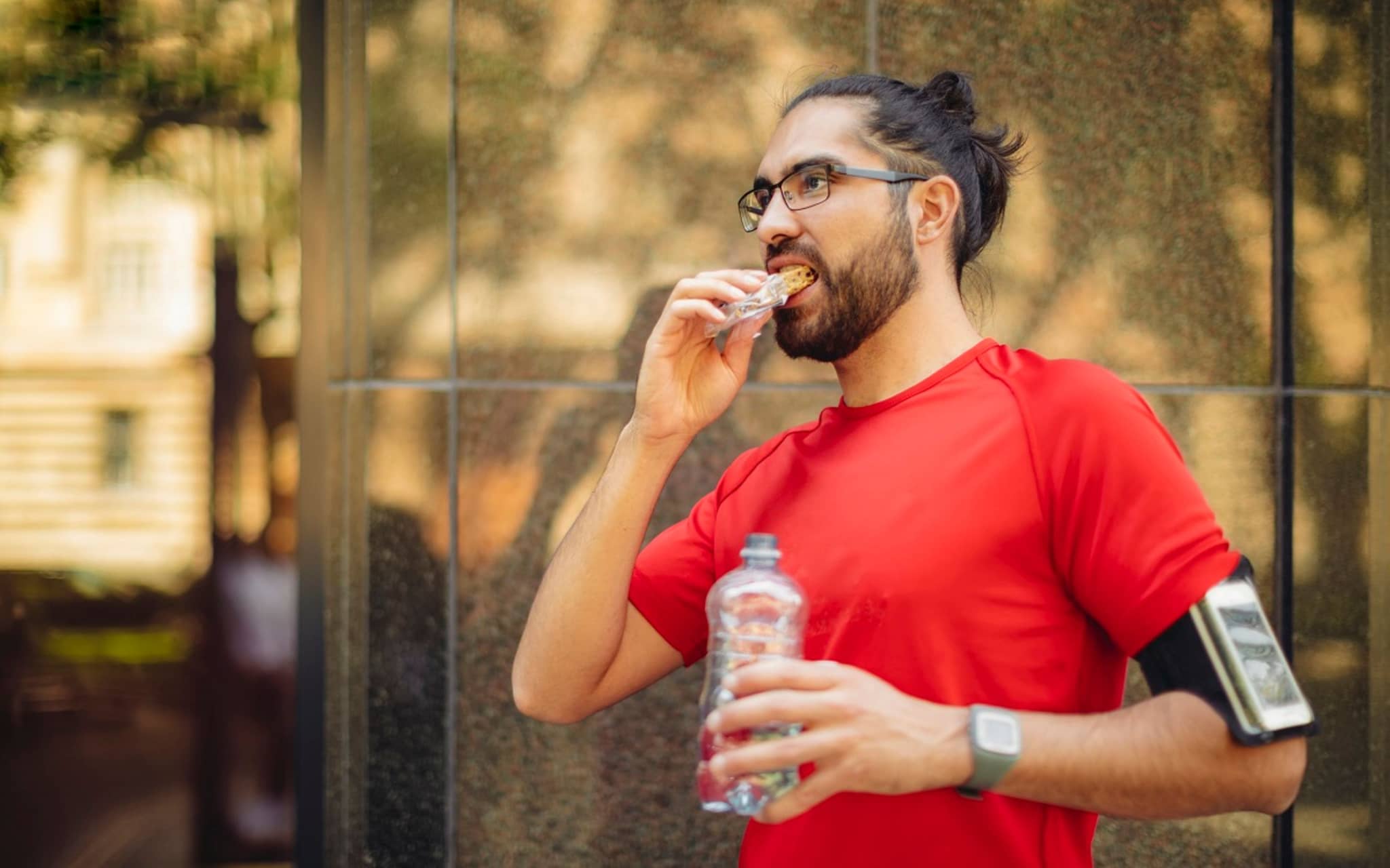 Fruchtriegel nach dem Sport: Mann isst nach dem Joggen einen Snackriegel und hält eine Flasche Wasser in der Hand.