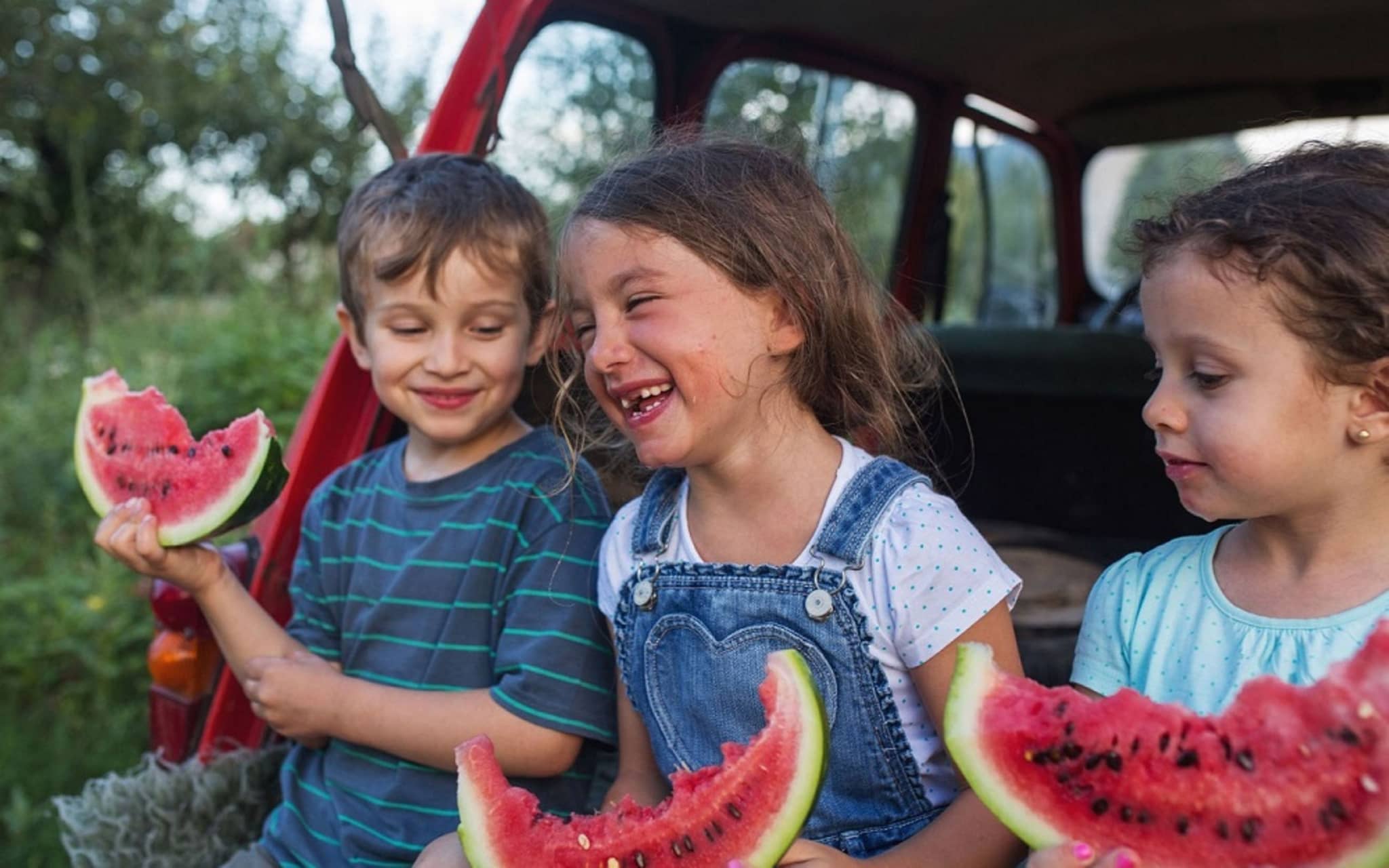 Safari-Kids: Drei Kinder sitzen lachend im offenen Kofferraum eines Autos und essen Melone.