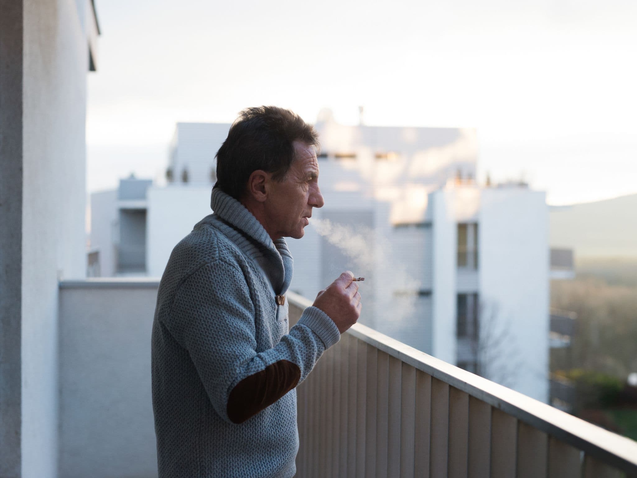 Älterer Mann steht nachdenklich auf dem Balkon und raucht dabei seine womöglich letzte Zigarette.