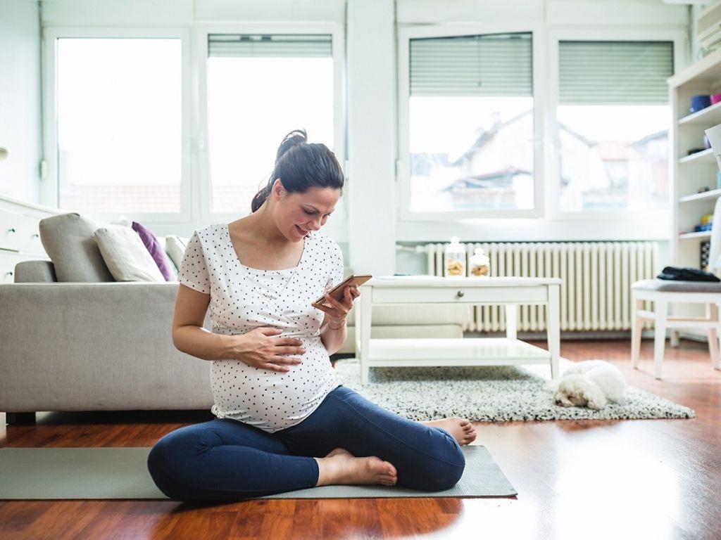 DAK Schwangerschaftscoaching: Schwangere sitzt auf dem Boden mit Handy in der Hand.