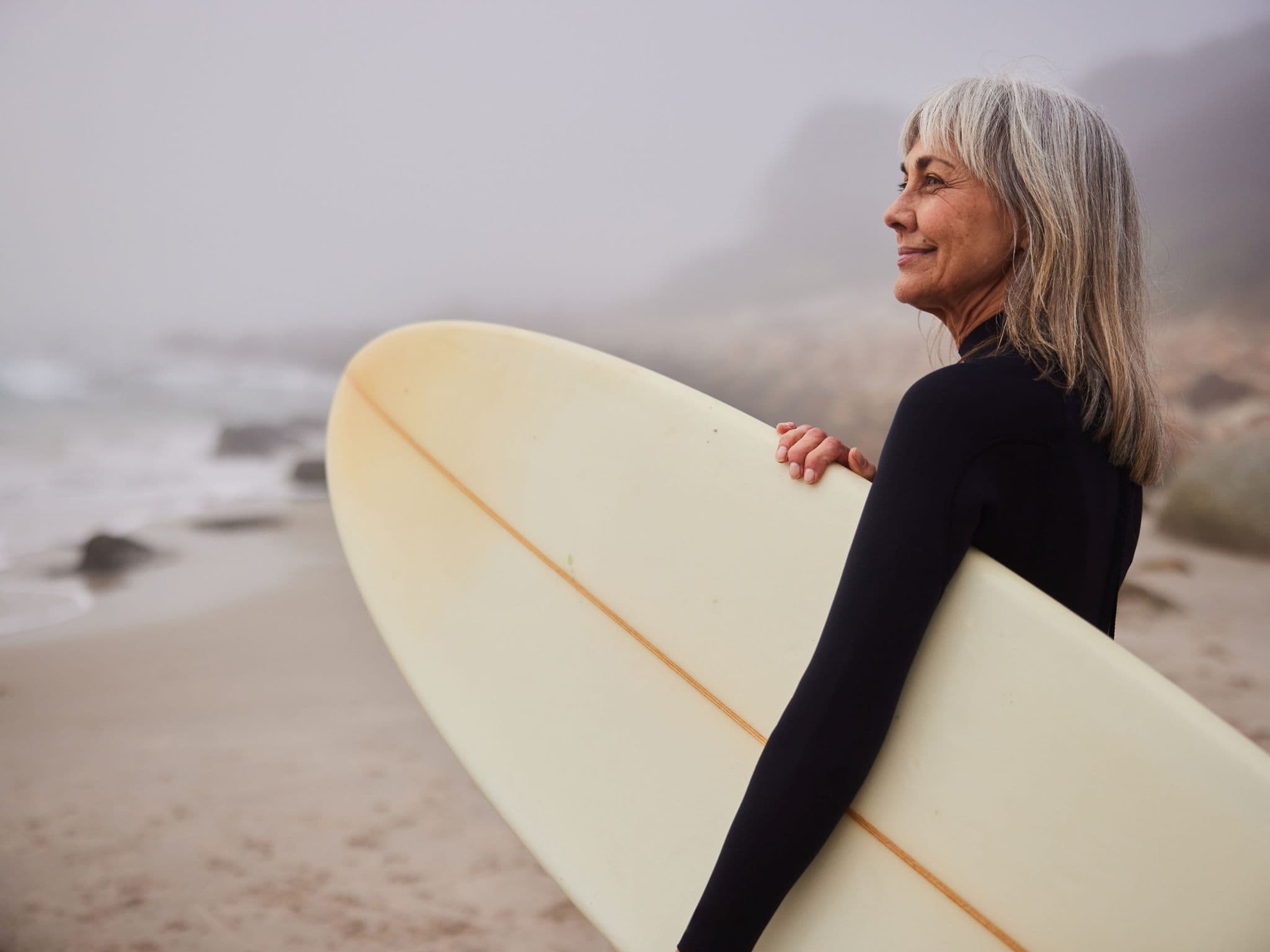 Postmenopause: Eine Frau mit grauen Haaren mit Surfbrett am Strand