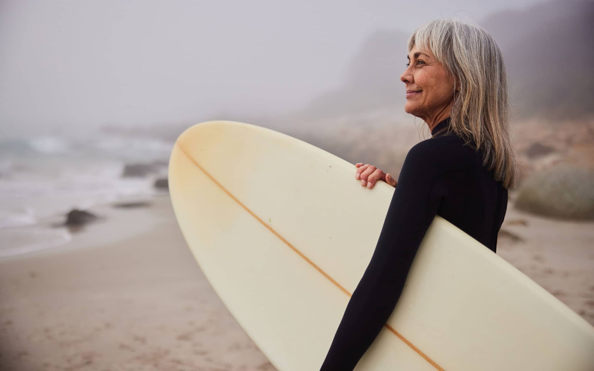Postmenopause: Eine Frau mit grauen Haaren mit Surfbrett am Strand