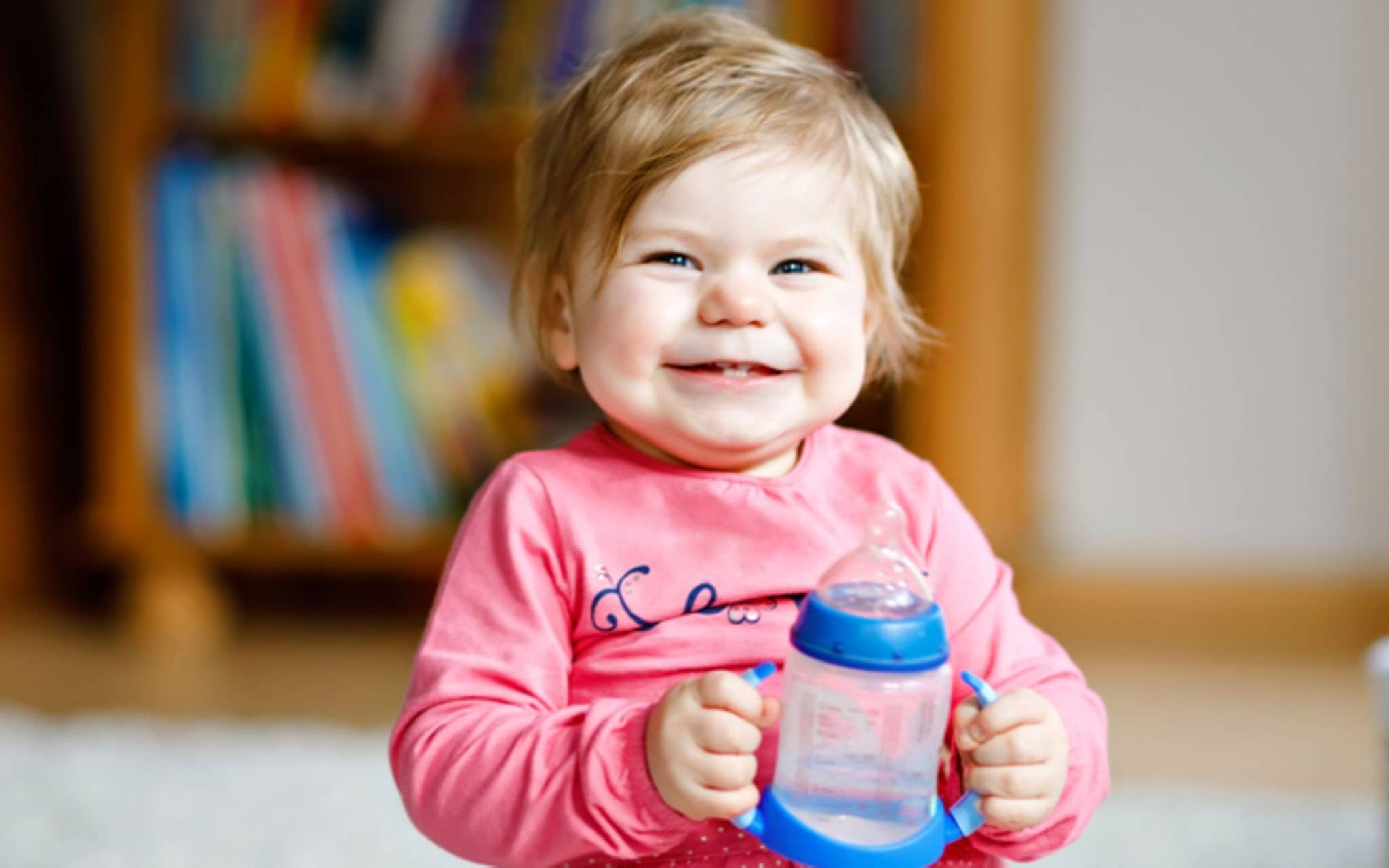 Babys trinken Wasser: Kleines Mädchen hält lachend eine Baby-Trinkflasche mit Wasser