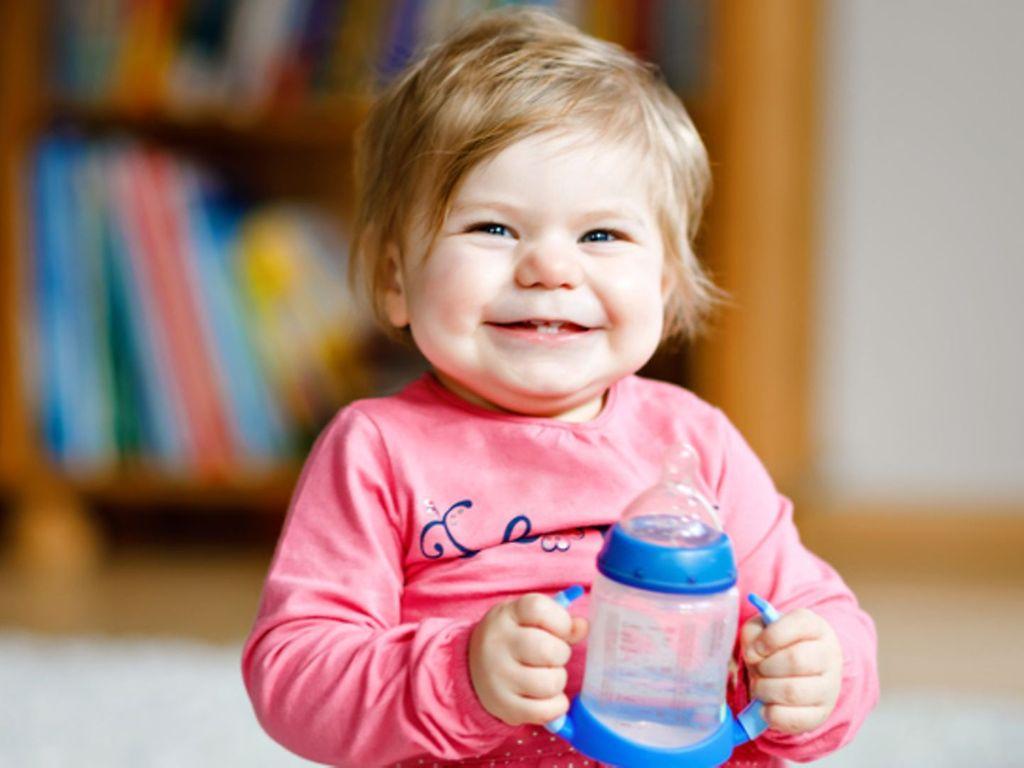 Babys trinken Wasser: Kleines Mädchen hält lachend eine Baby-Trinkflasche mit Wasser