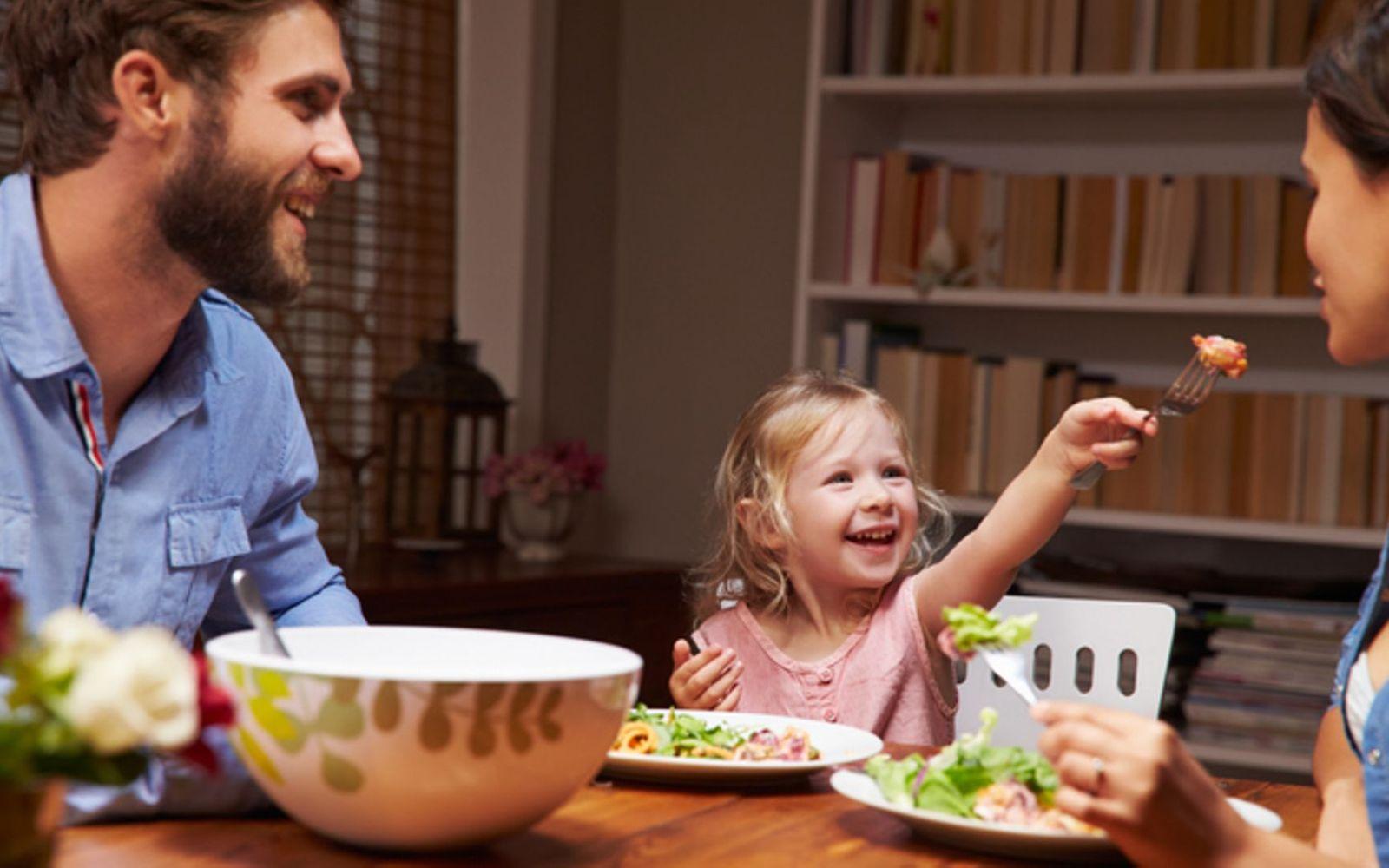 Ernährung für Kleinkinder: Junge Familie sitzt amüsiert am Esstisch und isst Salat
