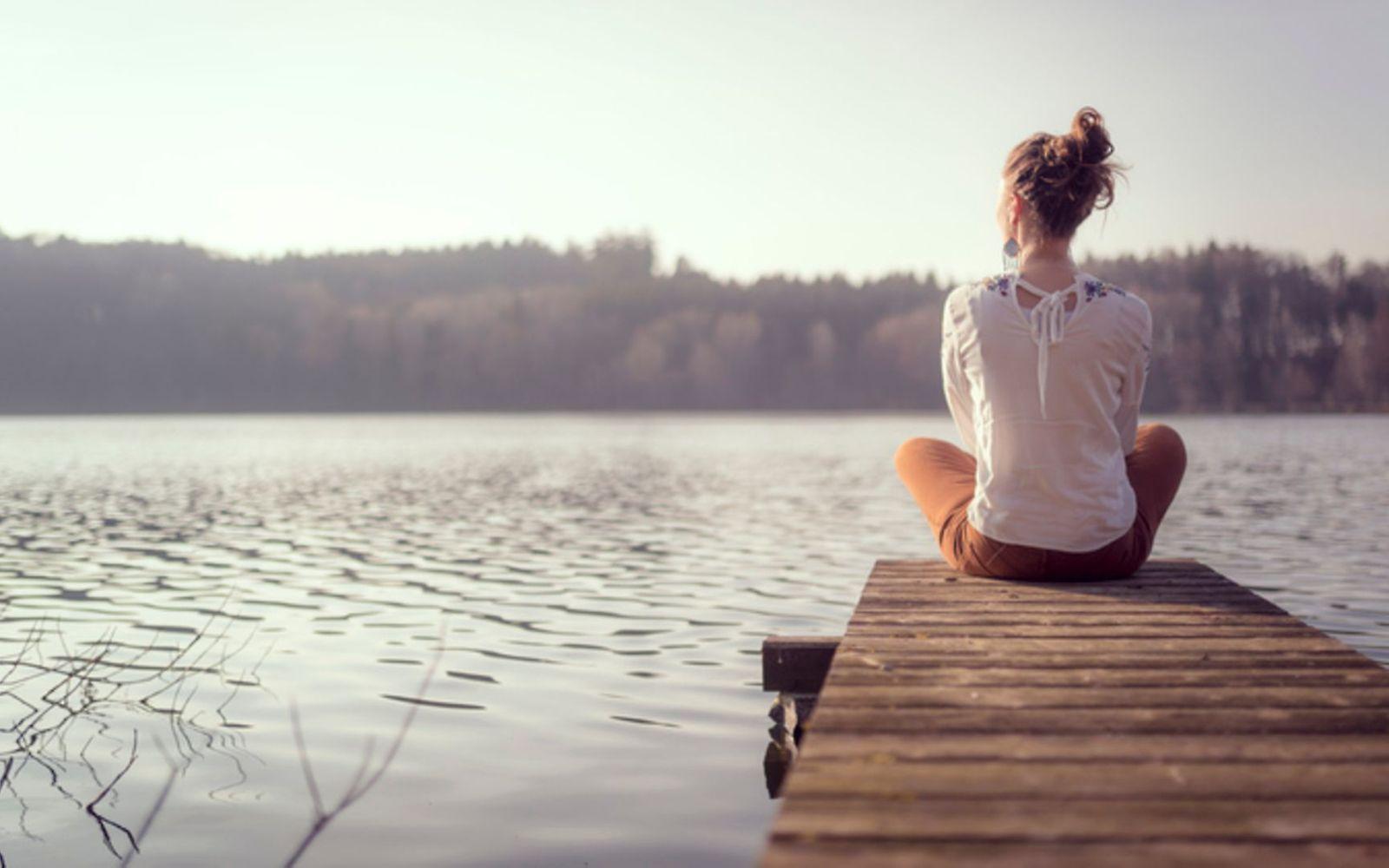 Hochsensibilität: Eine junge Frau sitzt im Schneidersitz auf einem Steg über einem See und genießt den Blick in die Natur