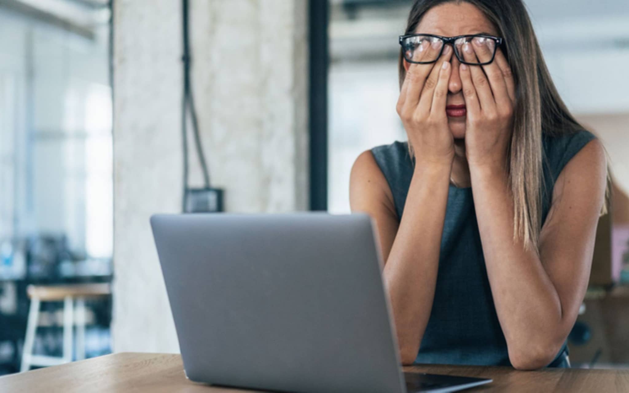 Augenringe: Frau mit Brille reibt sich die Augen, während sie am Schreibtisch vor einem Laptop sitzt