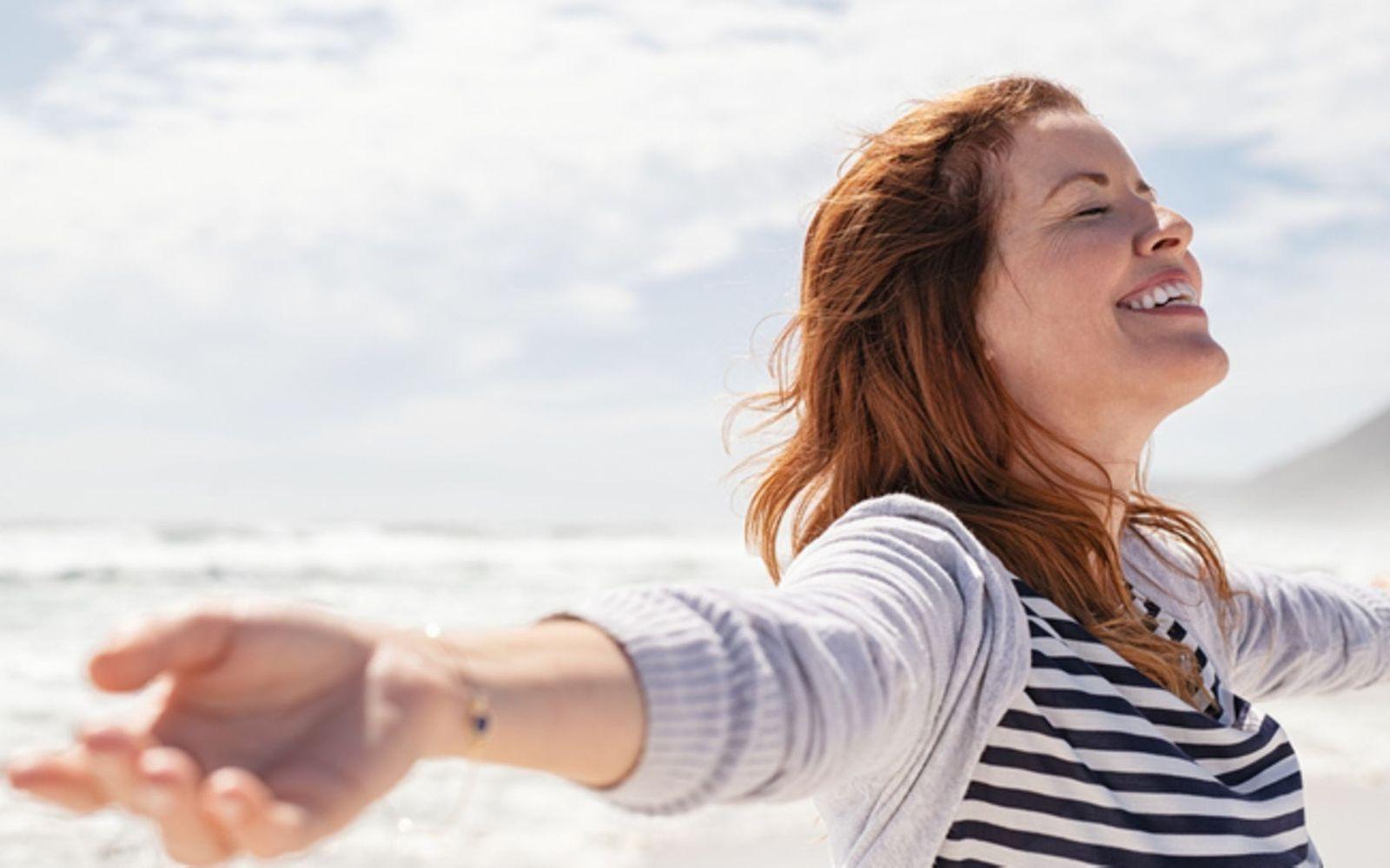 Kraft der Affirmationen: Frau steht lächelnd am Meer und genießt die Sonne und den Wind.
