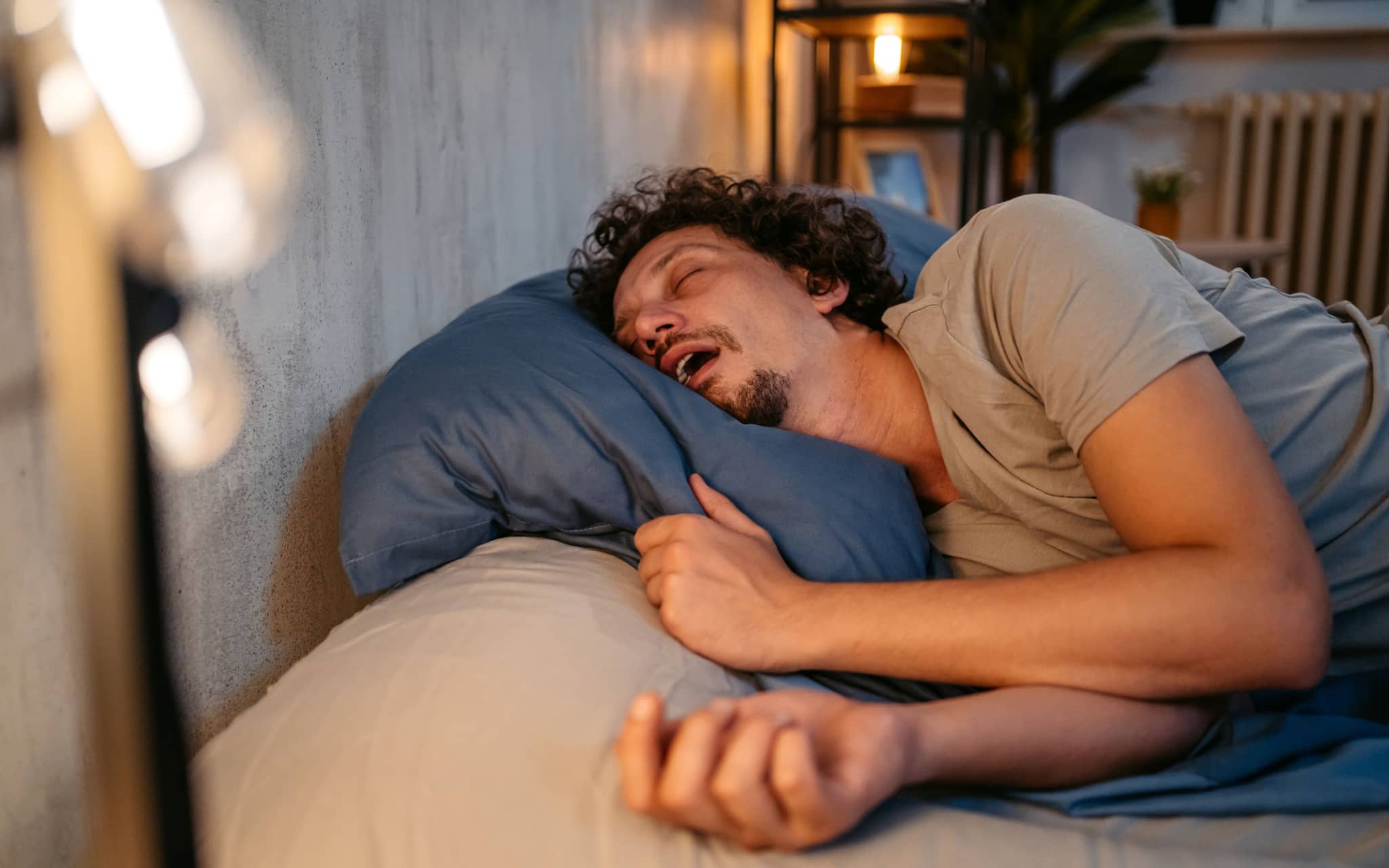 Ein Mann schläft mit weit geöffnetem Mund im Bett und scheint zu schnarchen.