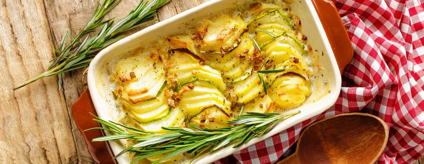 Rezeptfoto: Auflaufform mit Kartoffelgratin - daneben weiß-rot-karierteres Tuch und Holzlöffel.