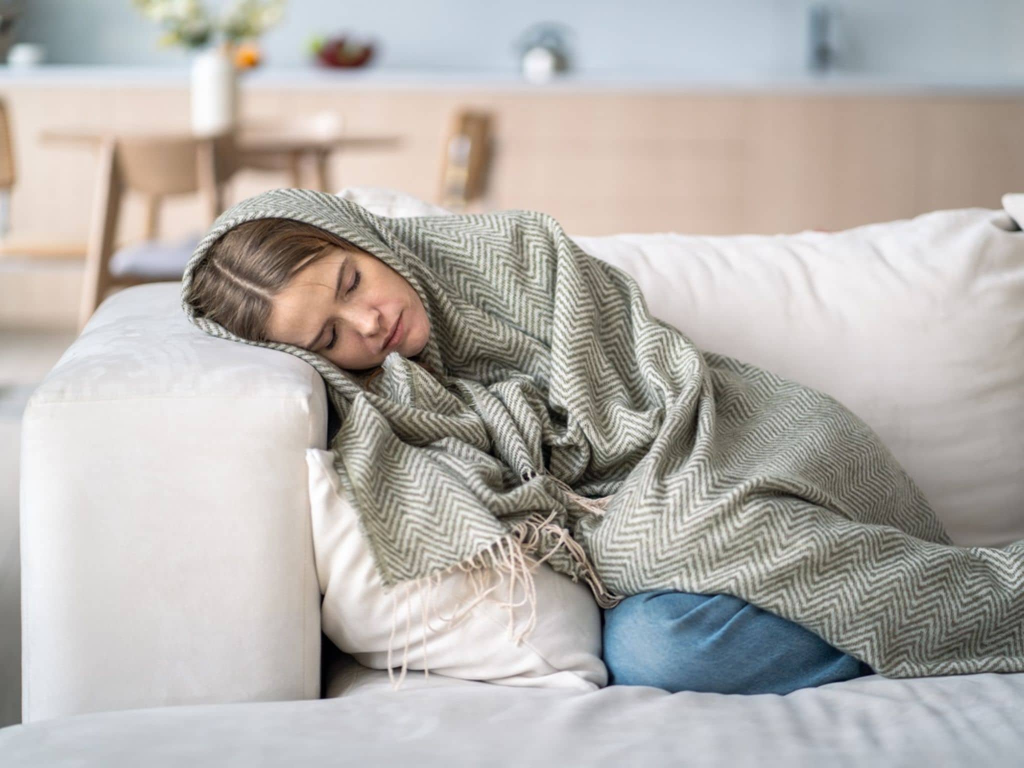 Anämie: Frau mit Blutarmut liegt in Decke eingewickelt mit geschlossenen Augen auf dem Sofa.