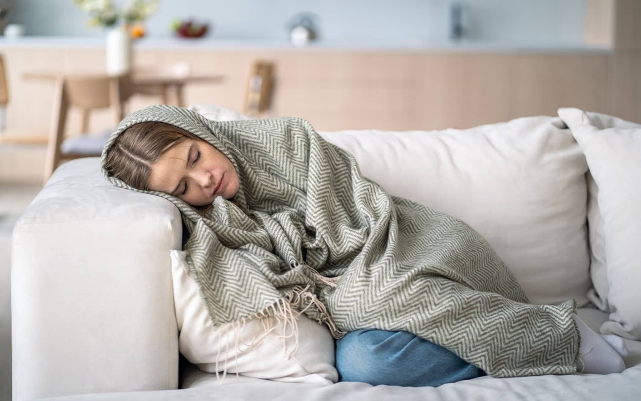 Anämie: Frau mit Blutarmut liegt in Decke eingewickelt mit geschlossenen Augen auf dem Sofa.