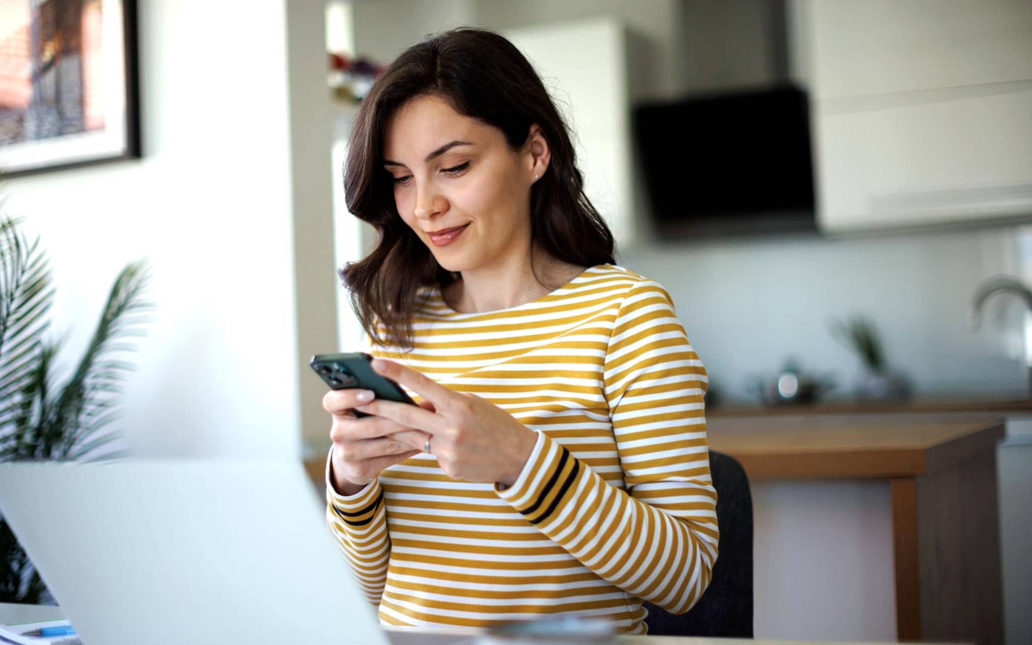 Lob & Beschwerde: Junge Frau sitzt zuhause am Tisch und tippt auf ihrem Smartphone.