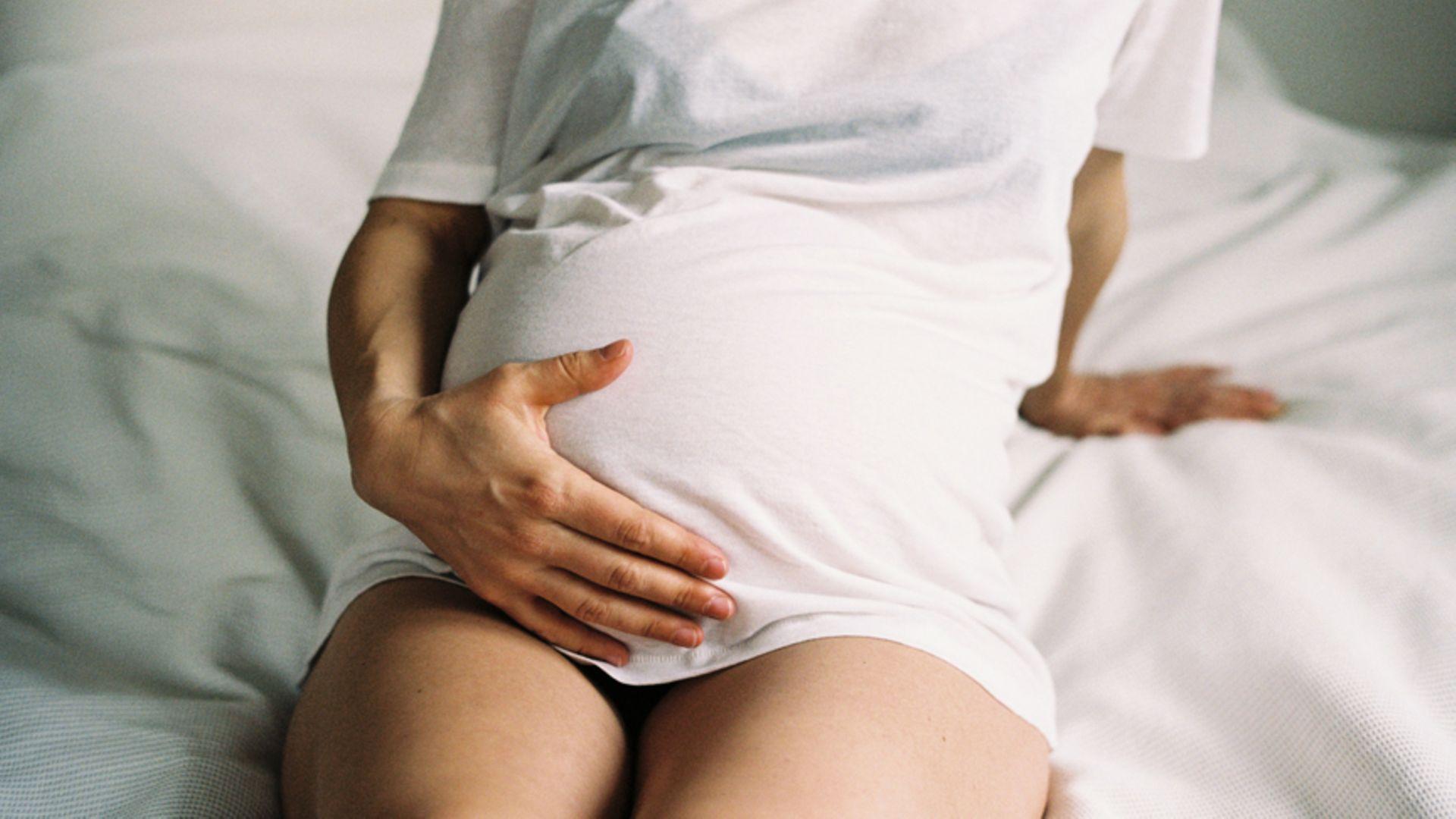 Chlamydien in der Schwangerschaft: Schwangere junge Frau, die auf dem Bett sitzt und ihren Bauch hält.