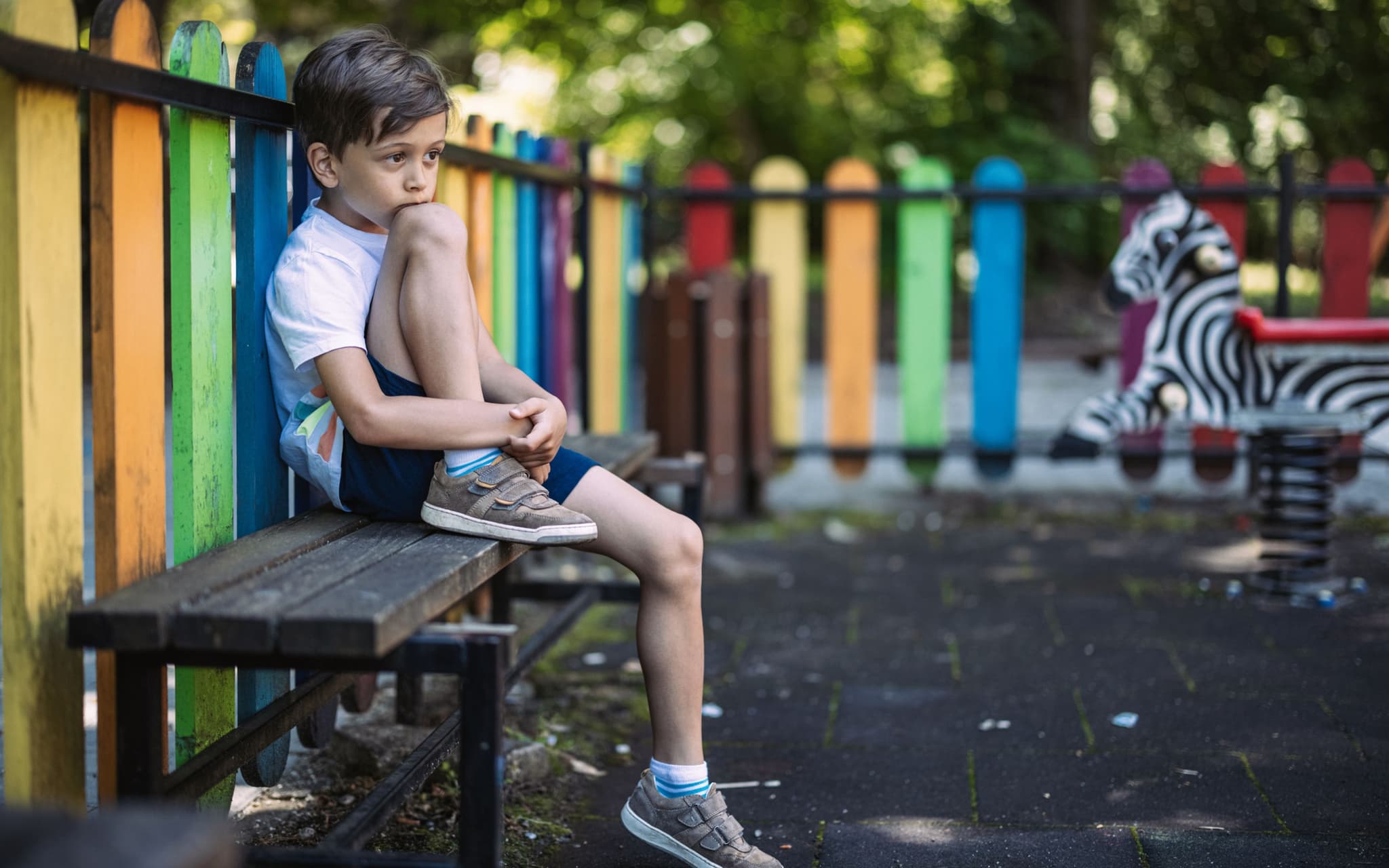 Psychische Erkrankungen bei Kindern: Ein Junge sitzt auf einer Bank am Randes des Spielplatzes und guckt traurig ins Leere
